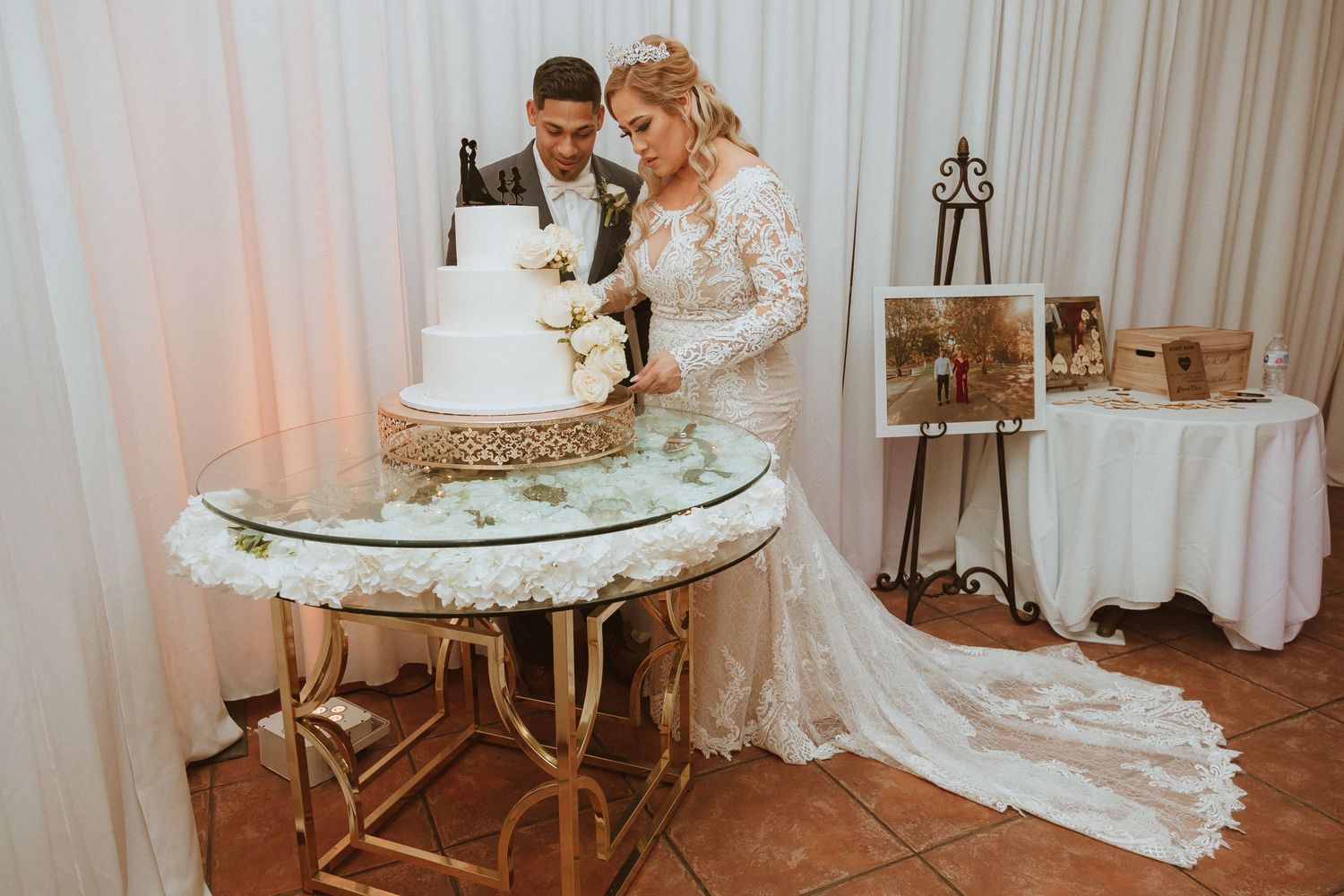 A newlywed couple cuts into their elegant white wedding cake on a gold-trimmed table during their reception.