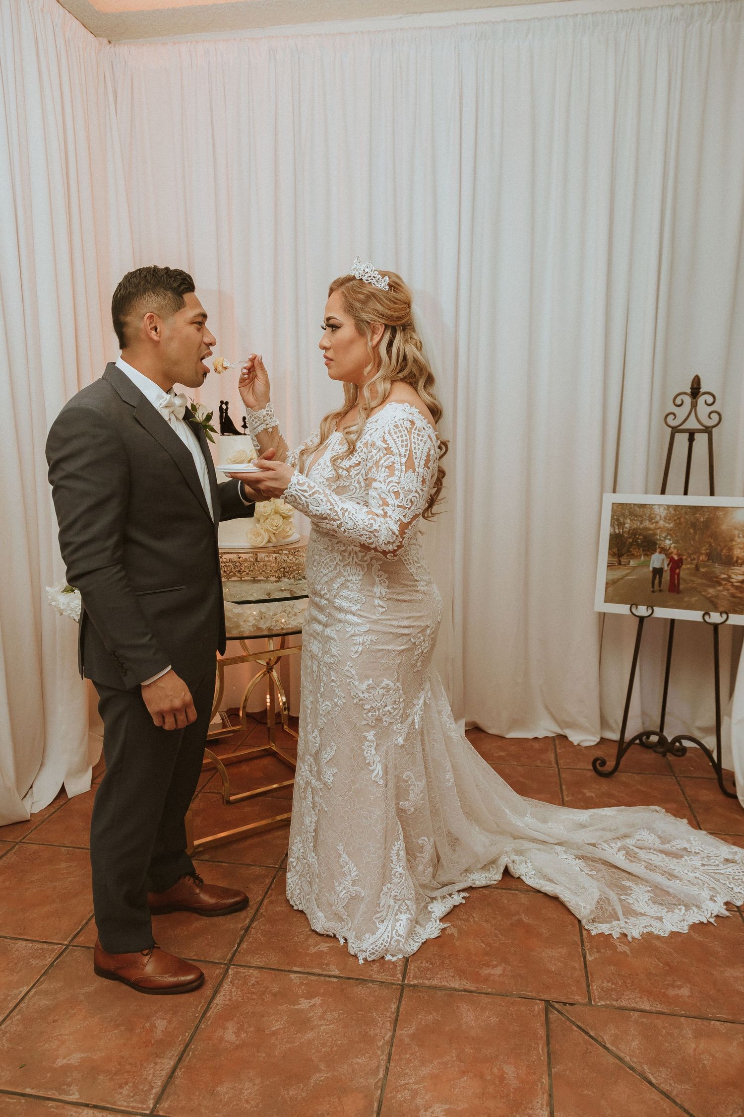 Newlywed couple tasting their wedding cake together in front of a white backdrop and terracotta tile floor.