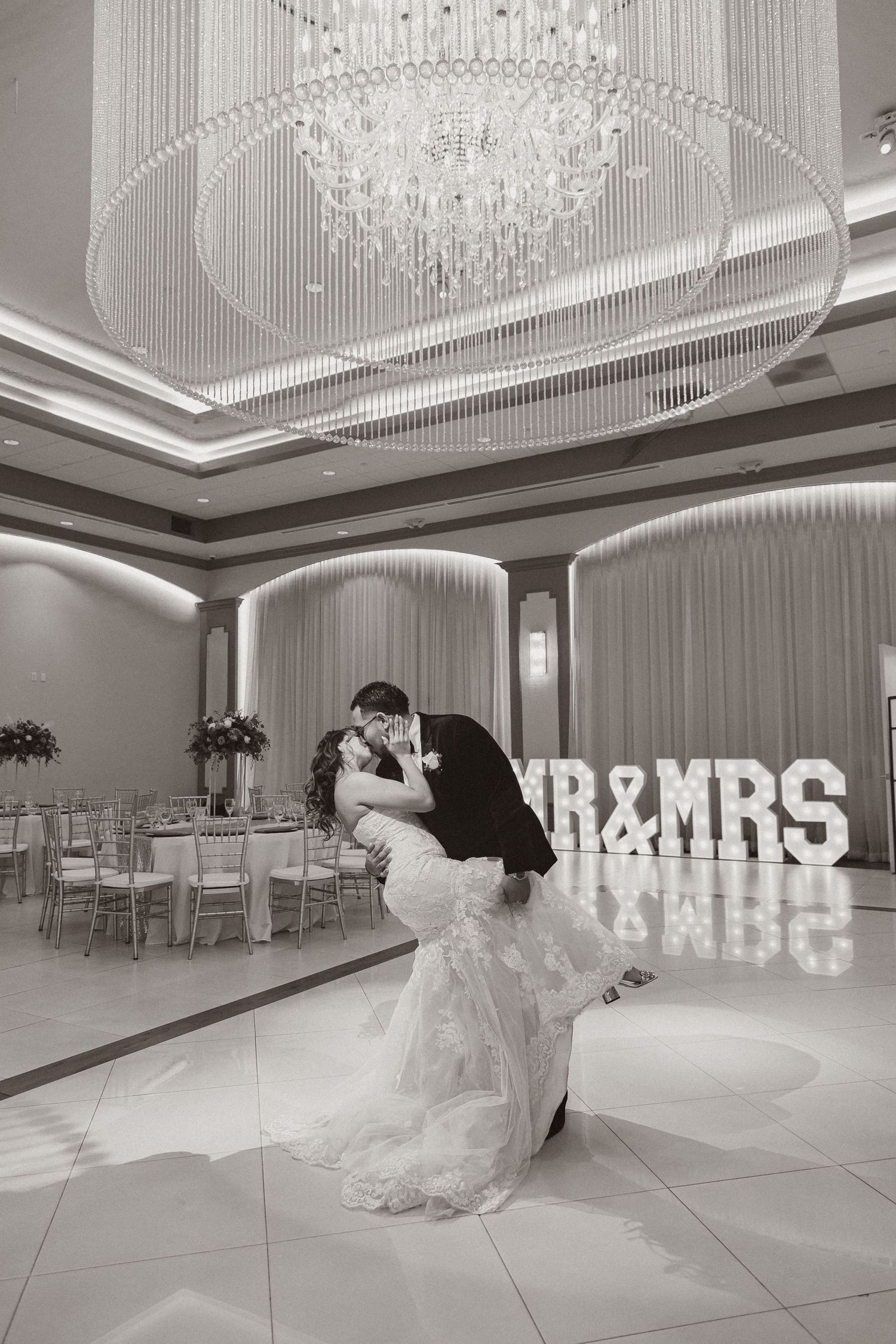 Black and white wedding portrait shows couple in dramatic dip pose under crystal chandelier.