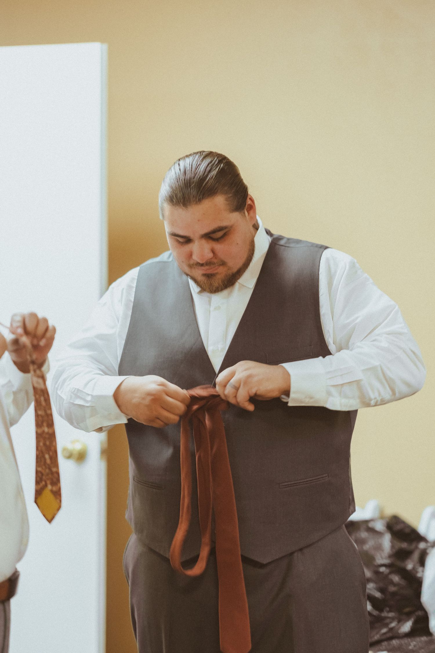 A groomsman adjusts a brown tie while getting ready in formal attire with a grey vest and white shirt.
