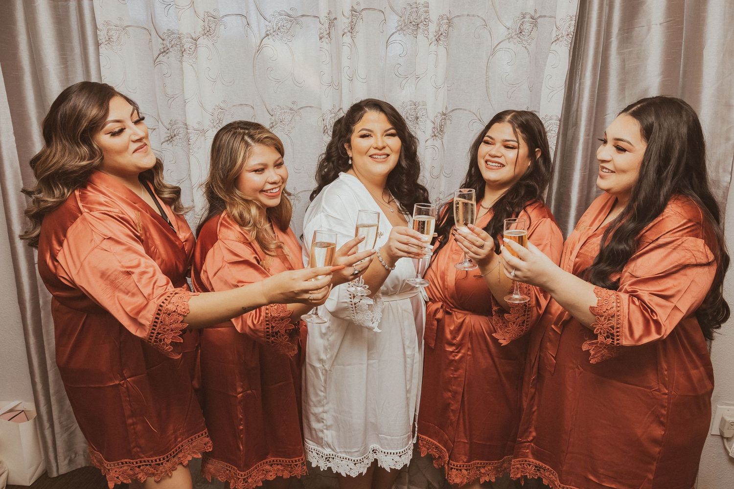 Bridal party in matching orange robes toasts with champagne glasses against a light colored backdrop.