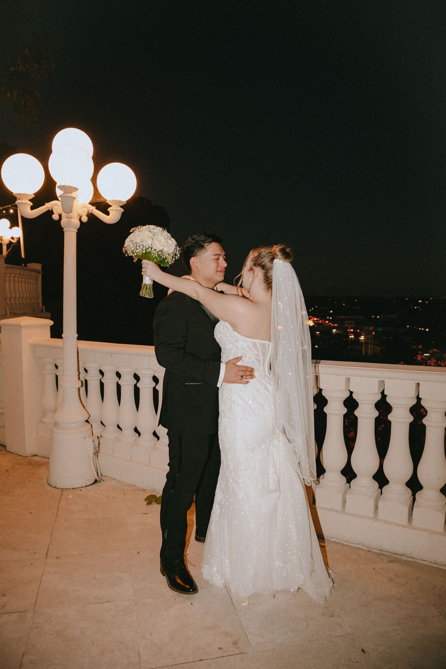 A newlywed couple embraces during their nighttime wedding photoshoot on an elegant balcony.