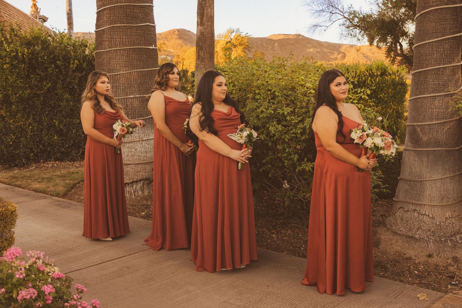 Four bridesmaids in burgundy dresses stand in a row beside palm trees.