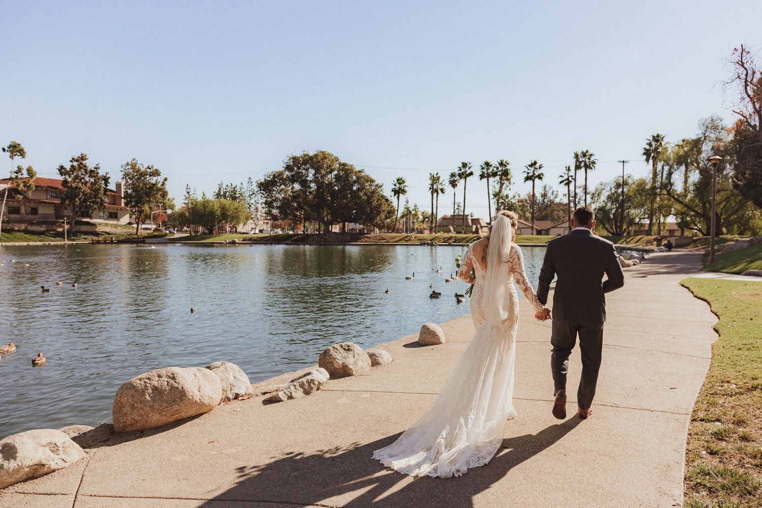 A couple in wedding attire walks along a lakeside path with palm trees in the background on their special day.