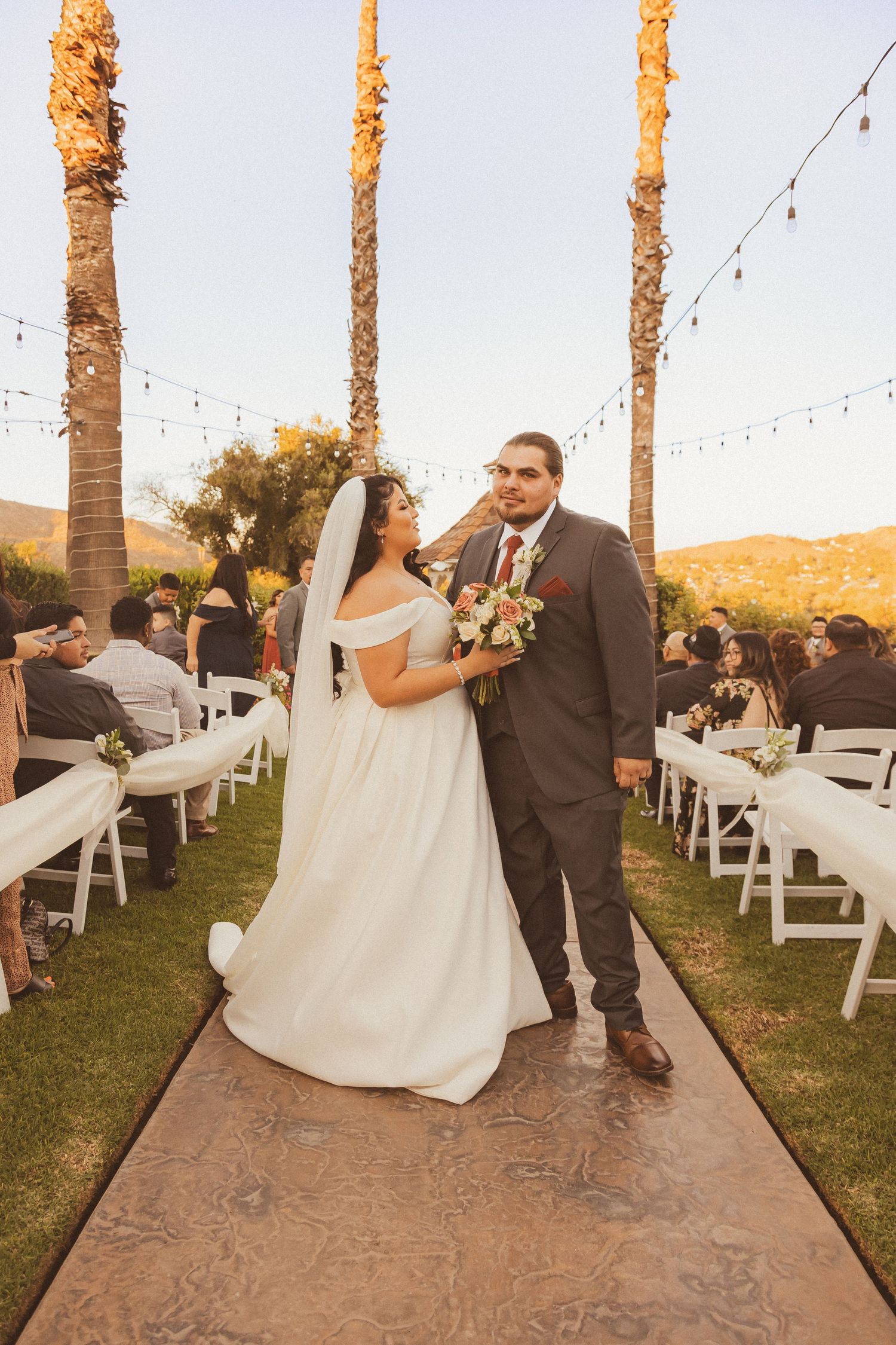 A couple poses during their outdoor wedding ceremony with palm trees and string lights in the background.