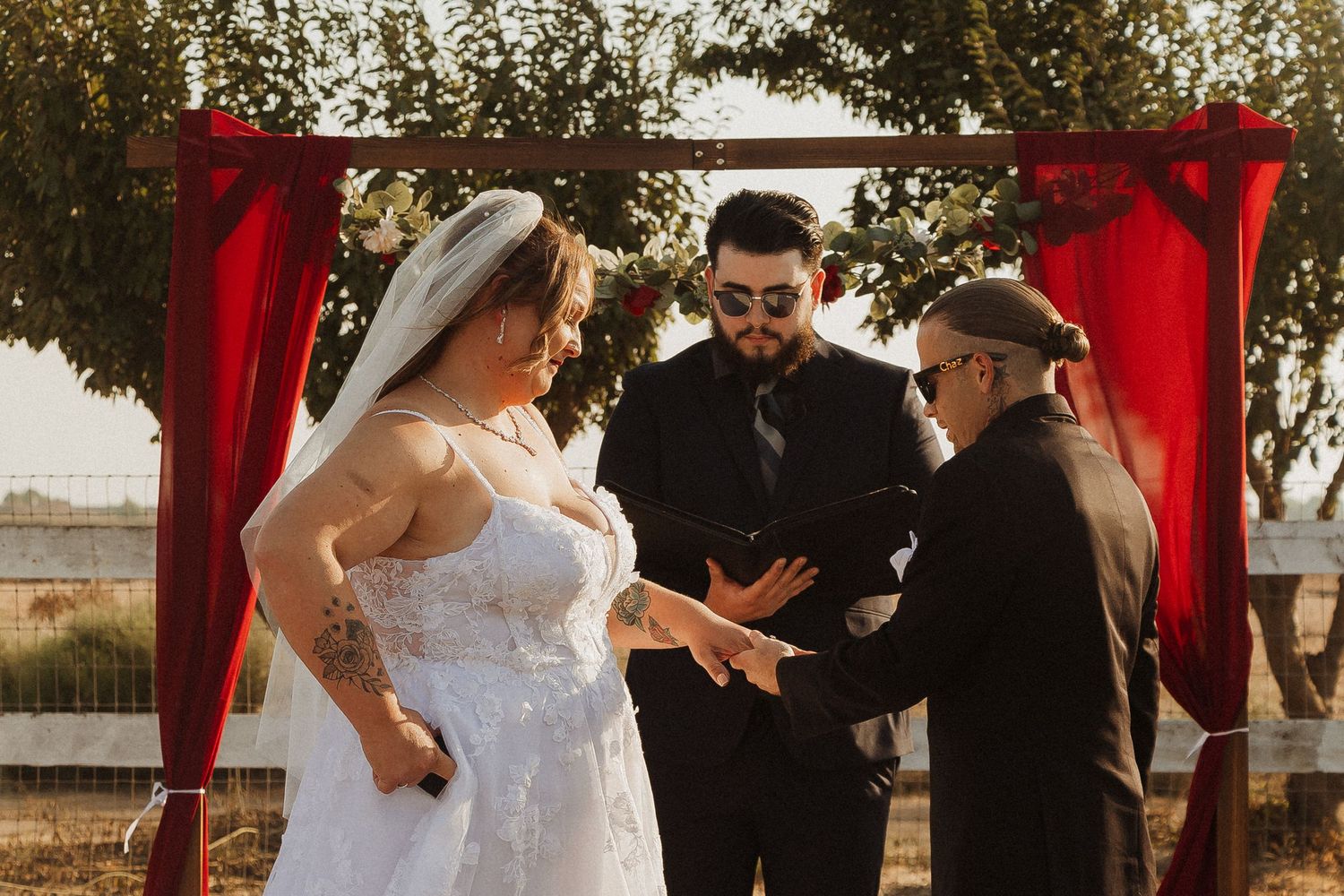 A wedding ceremony in progress under a red arch against a rural landscape.