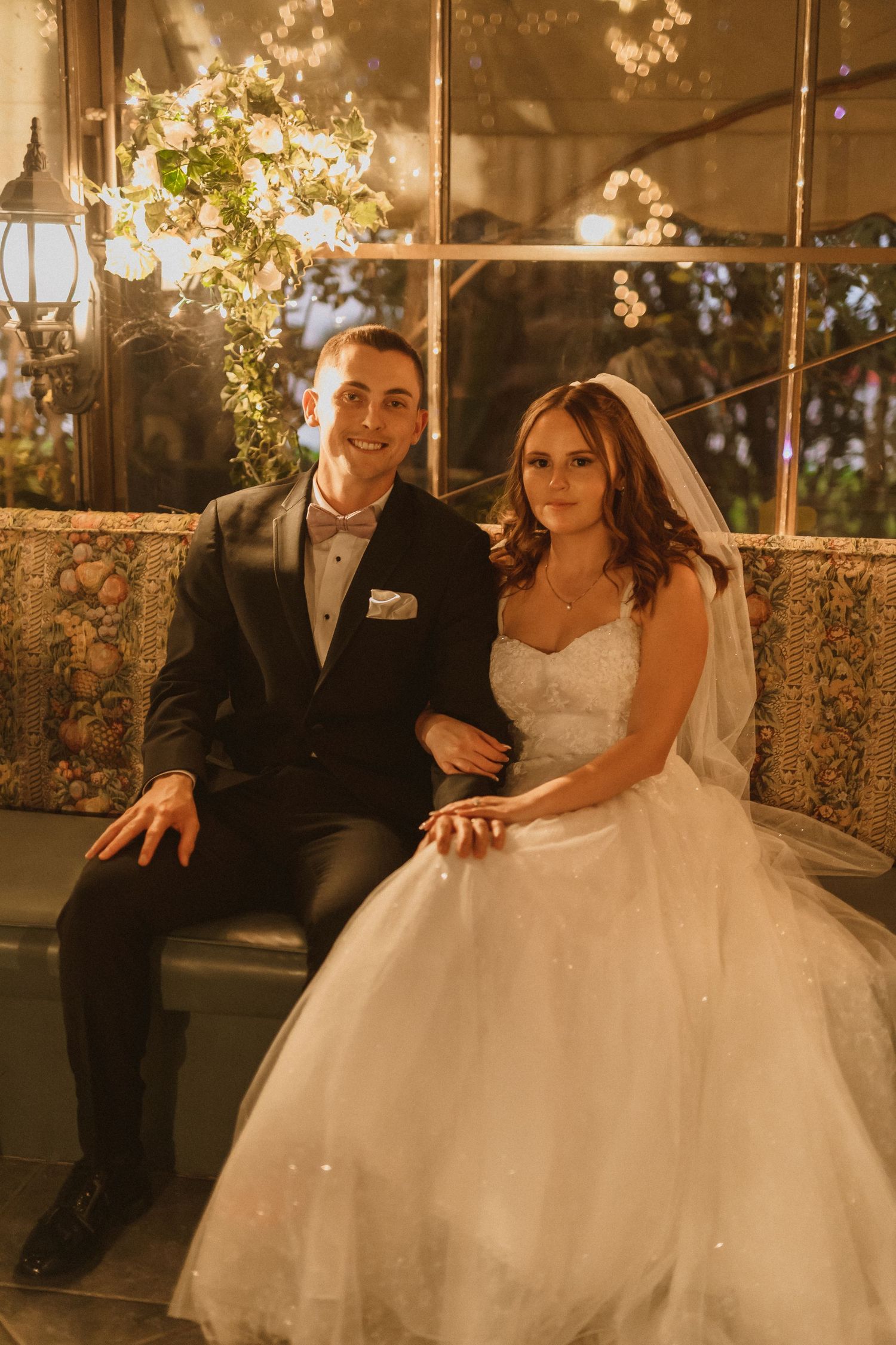 Newlyweds sit together under ornate floral lighting at their reception venue.