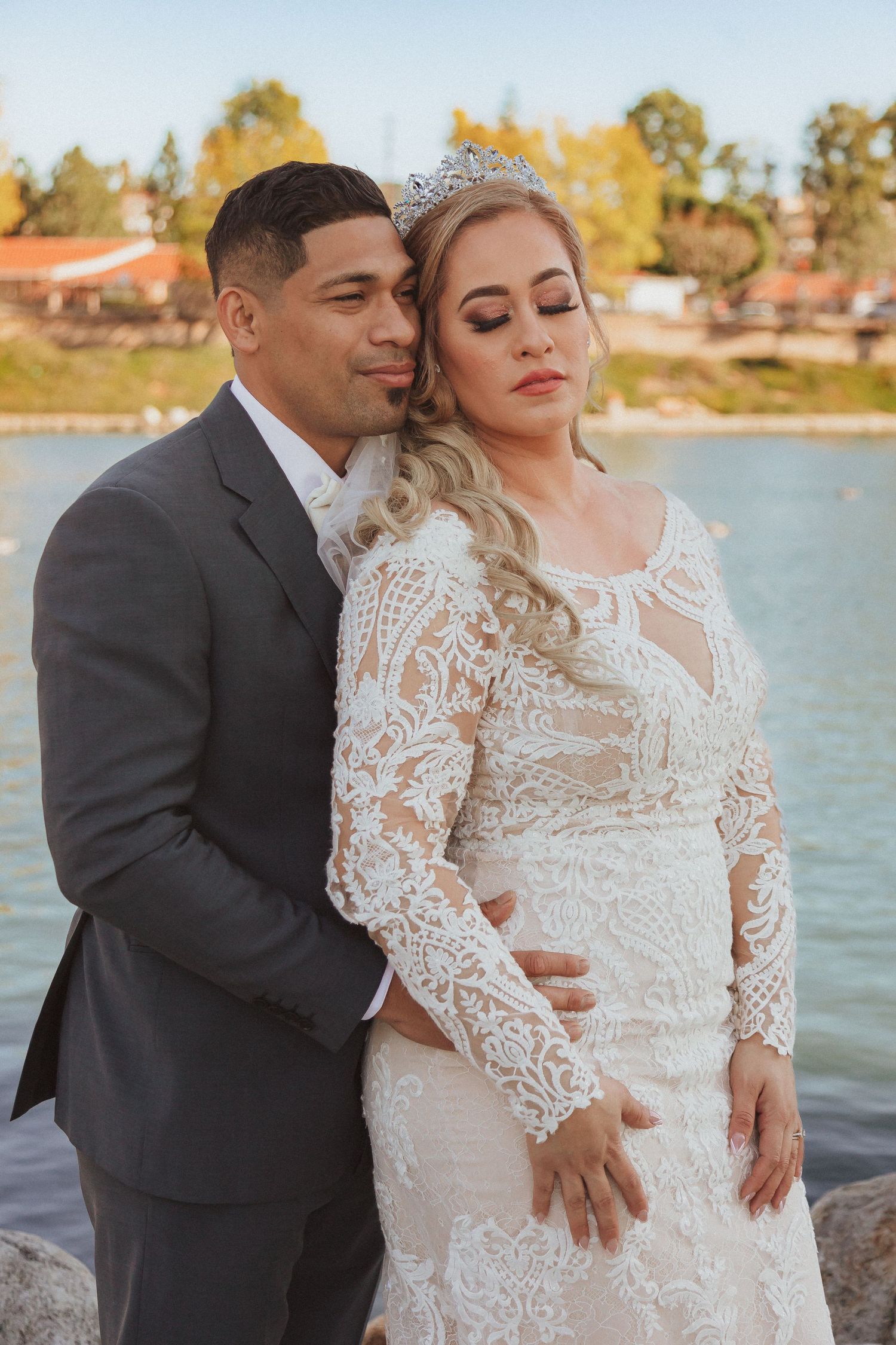 Elegant couple poses by lakeside in formal wedding attire during golden hour.