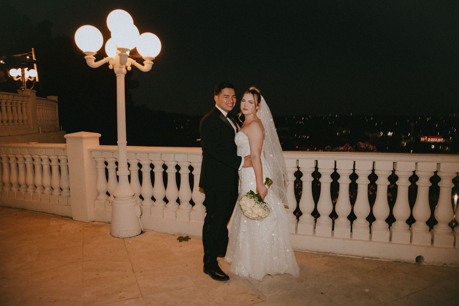 Nighttime wedding photo featuring couple on illuminated terrace with city backdrop.