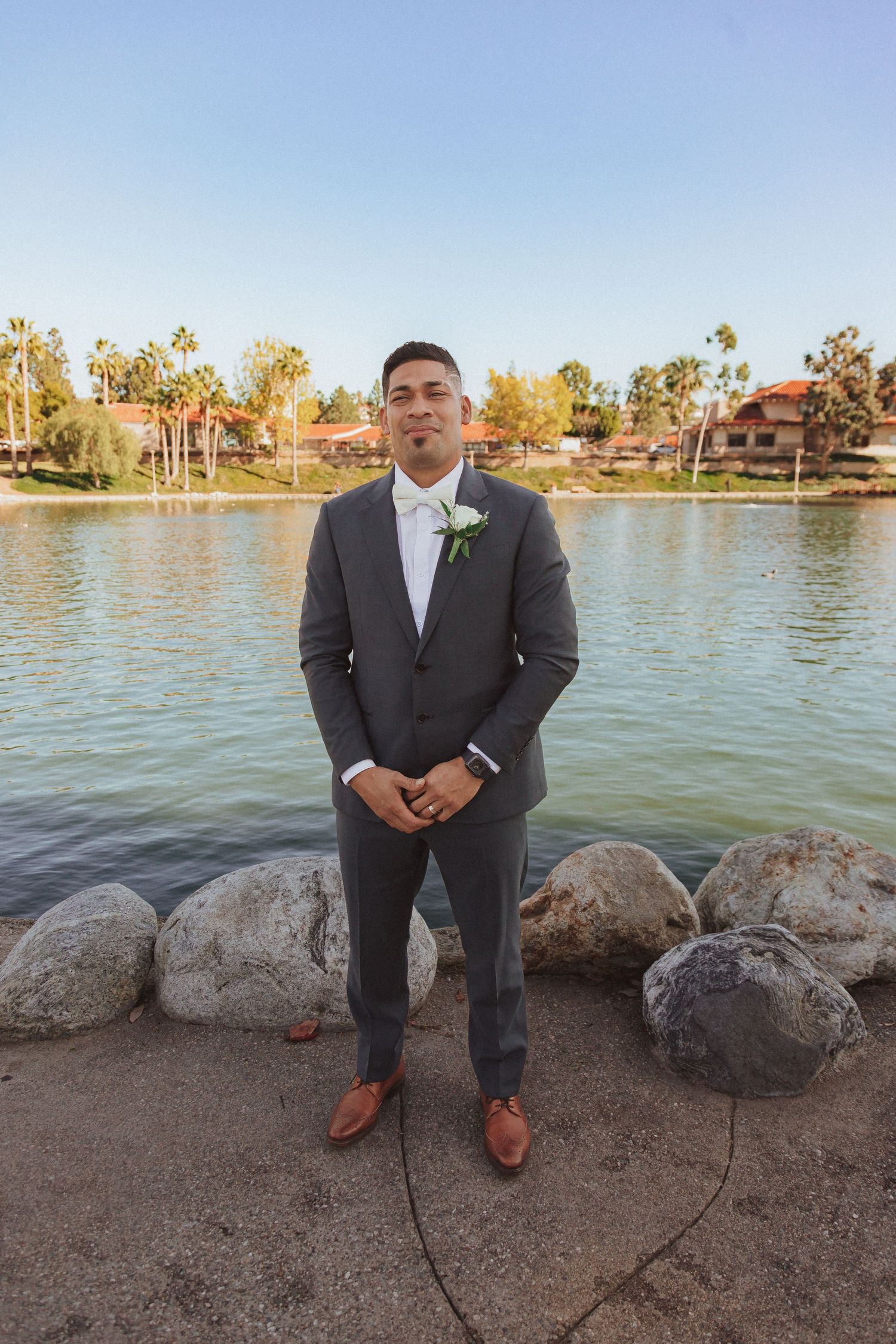 A groom in a charcoal suit poses by a lakeside location with palm trees and rocks in the background.