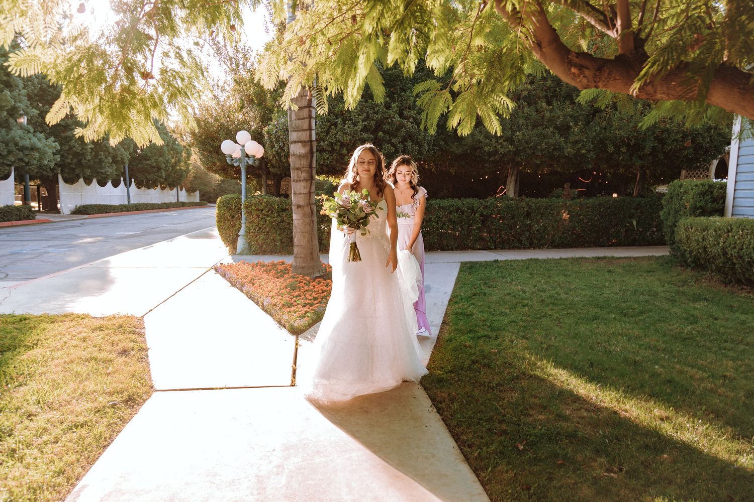 Bride in flowing white gown walks along sunny garden pathway.