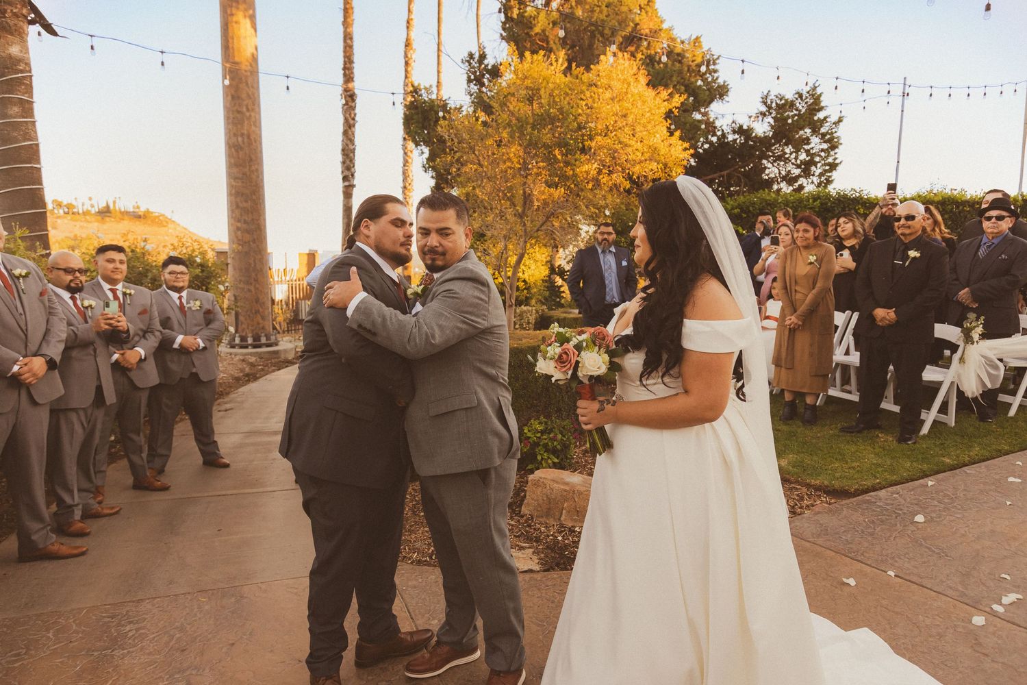 Wedding guests in gray suits share emotional moments during an outdoor ceremony.