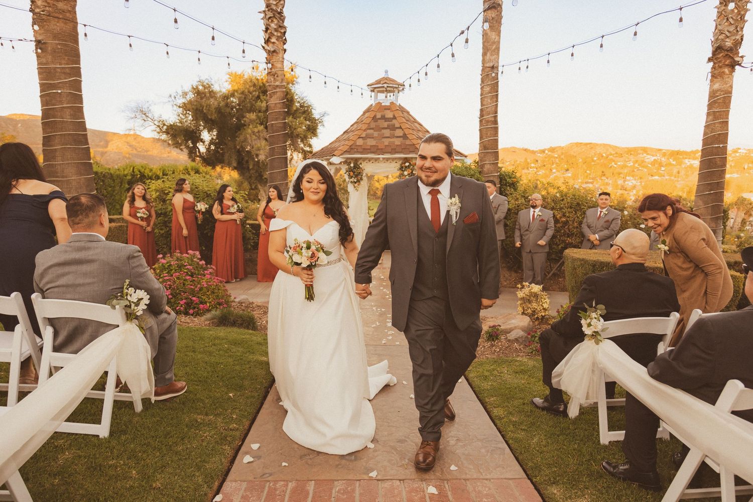 Wedding recessional along brick path with string lights and white chairs.