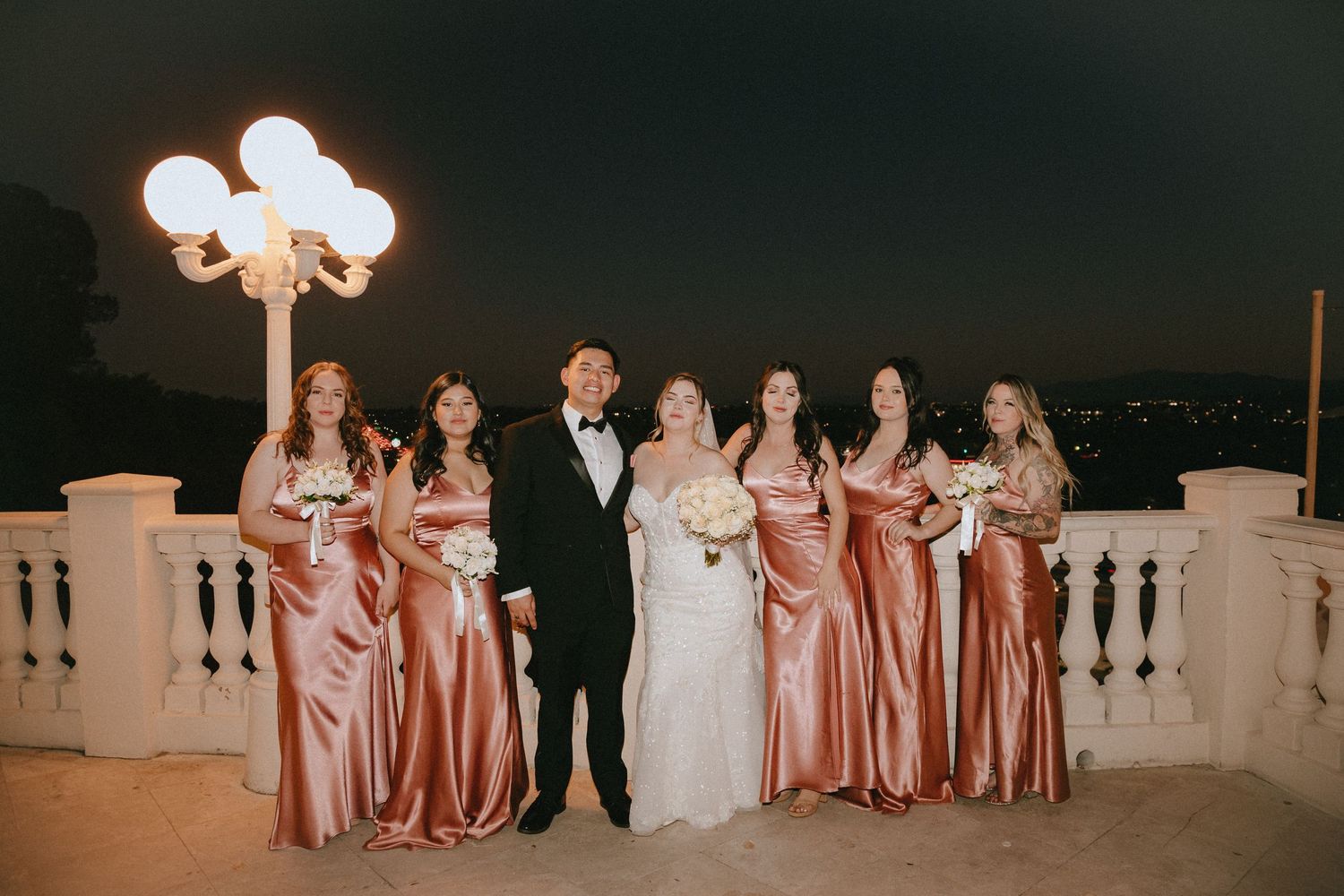 Wedding party in formal attire and blush satin dresses gathered on terrace under vintage lamp post.