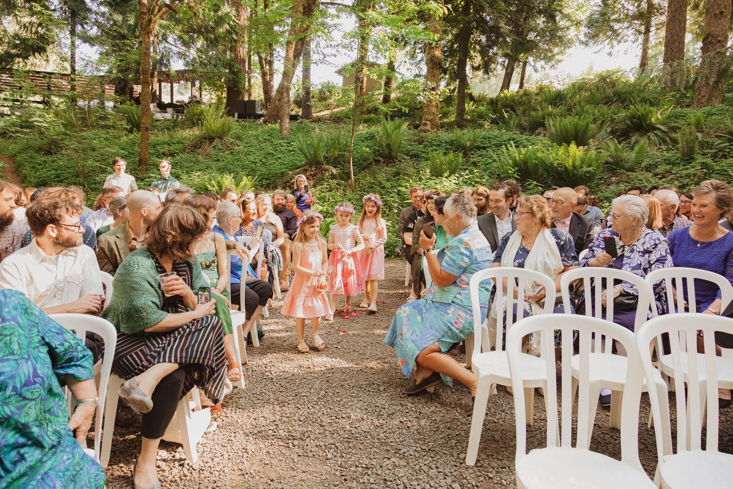 Outdoor forest wedding ceremony with guests seated on white chairs along a natural path.