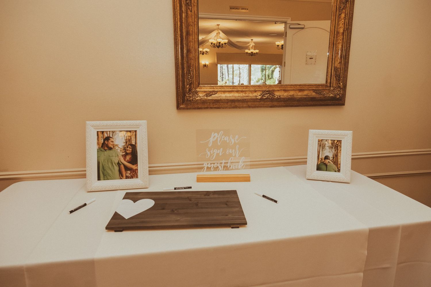 A wedding guest book table with framed photos and a wooden sign against a beige wall with mirror.