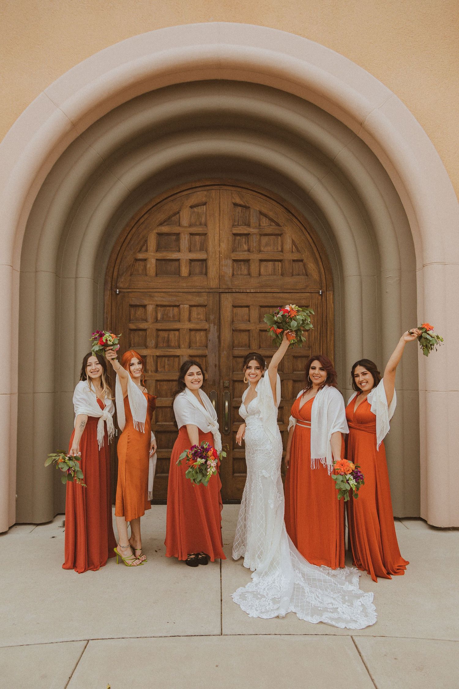 Wedding party wearing orange dresses and white shawls pose with bride in front of arched wooden doorway.