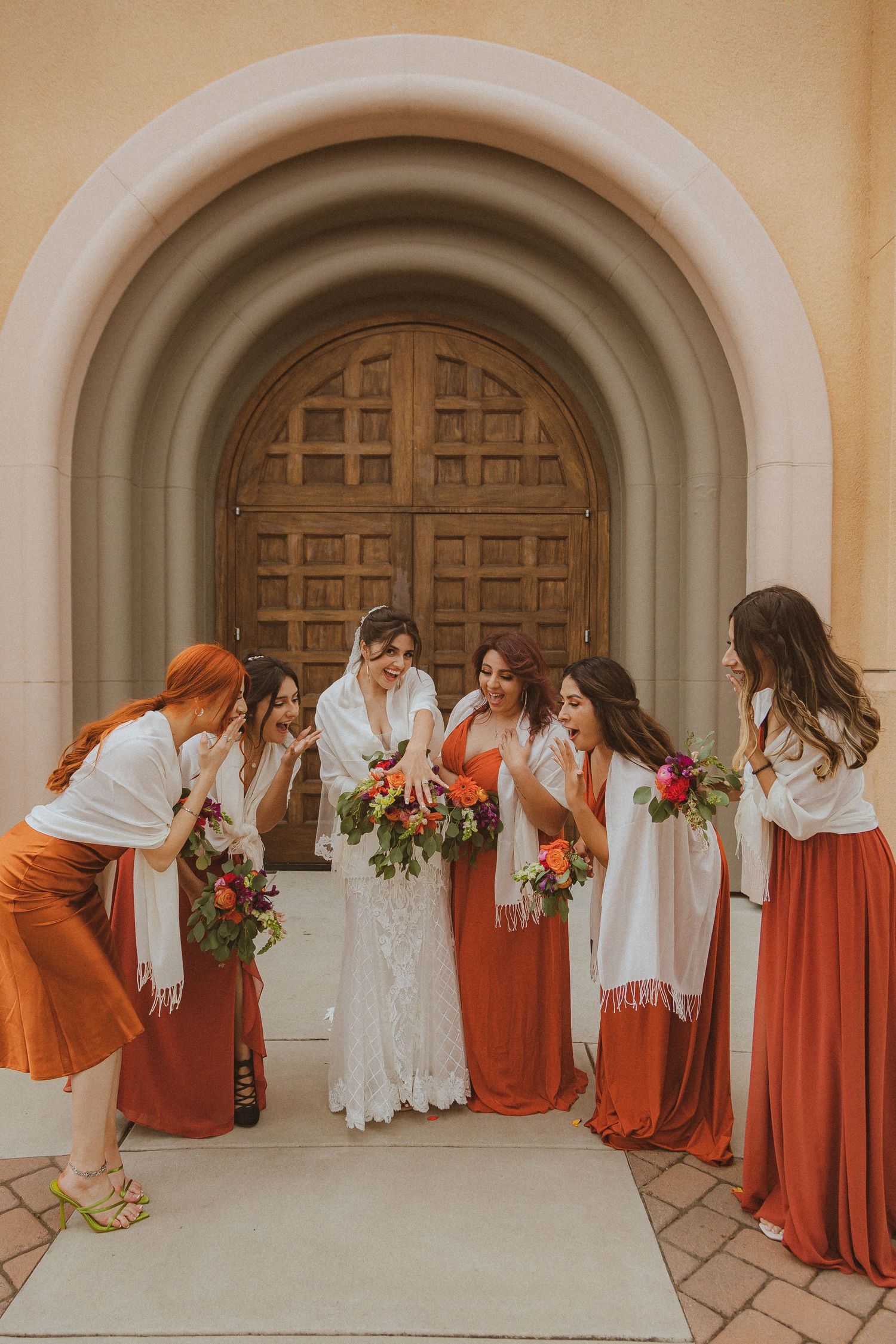 Wedding party in orange dresses shares a candid laughing moment with bride under arched doorway.