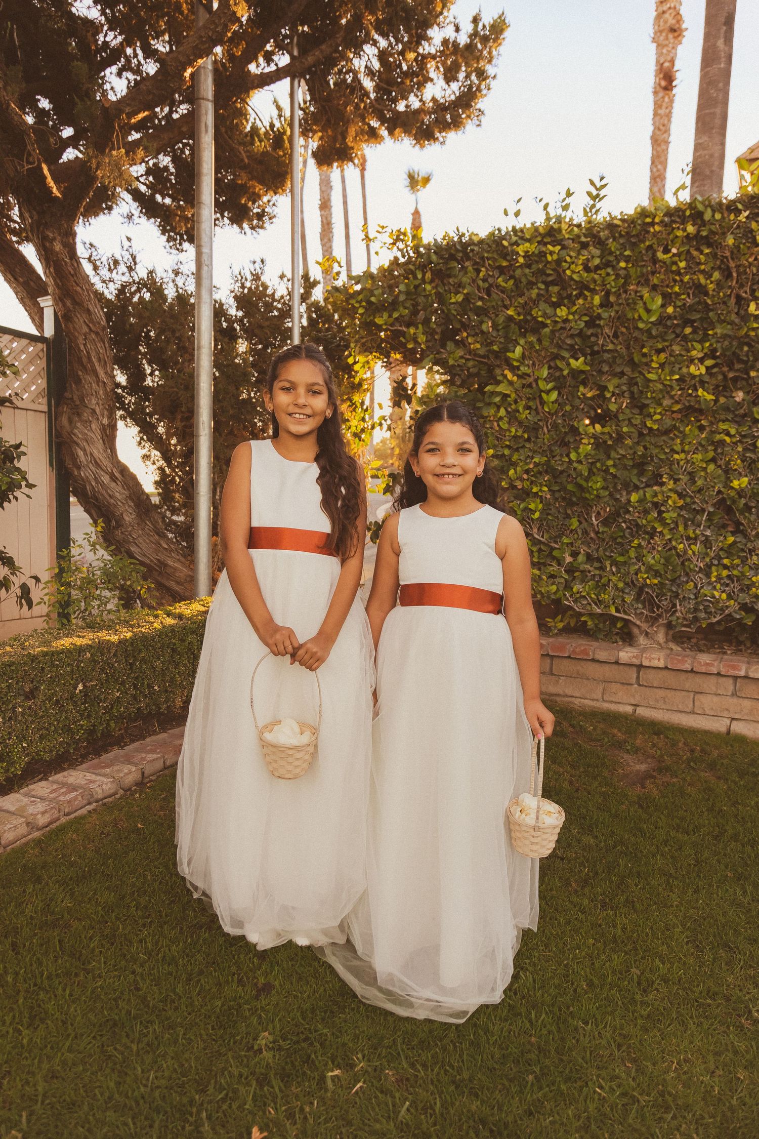 Two flower girls in white dresses with orange sashes hold baskets in a garden setting.