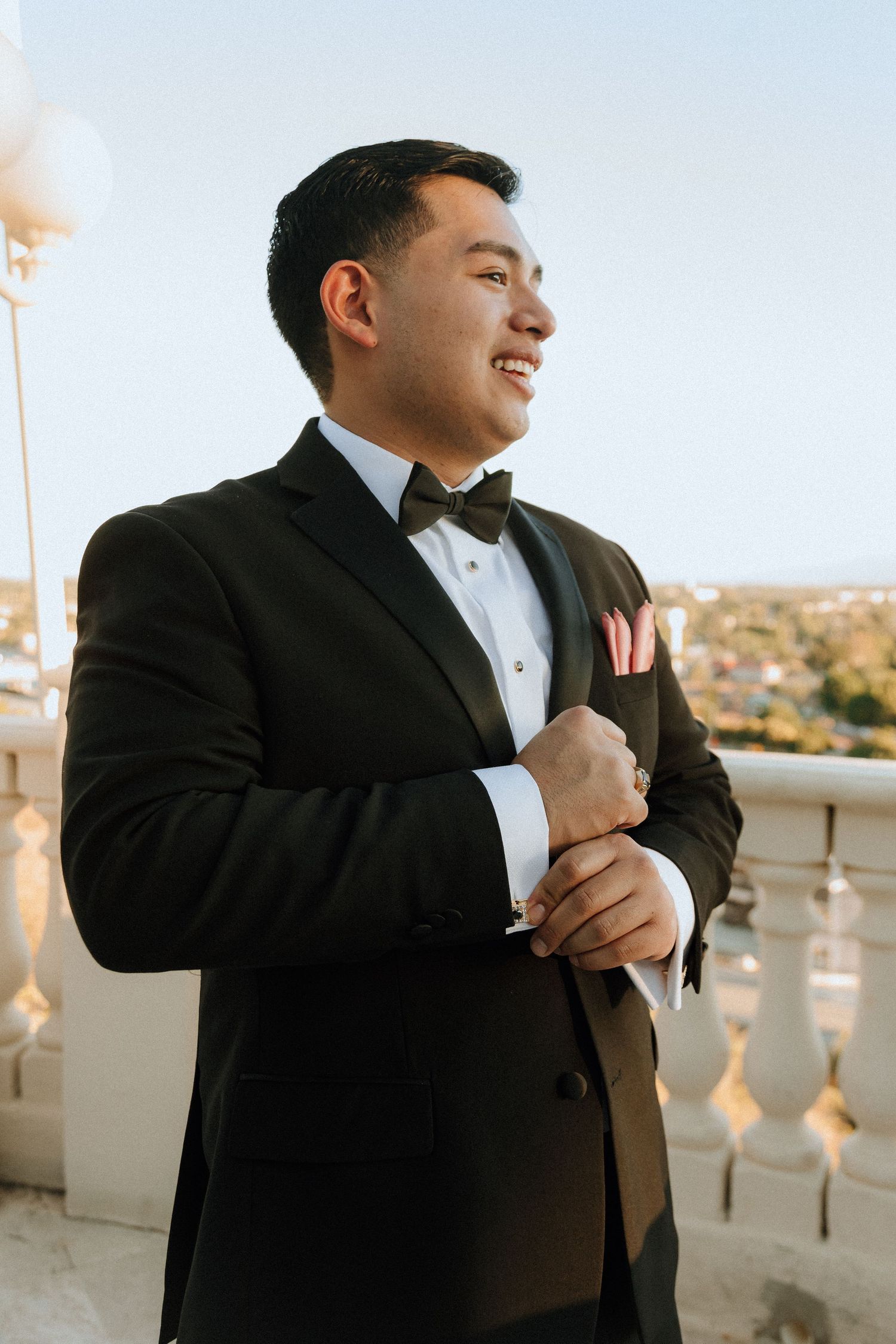 A groom in a classic black tuxedo and bowtie adjusts his cuff on an outdoor balcony.