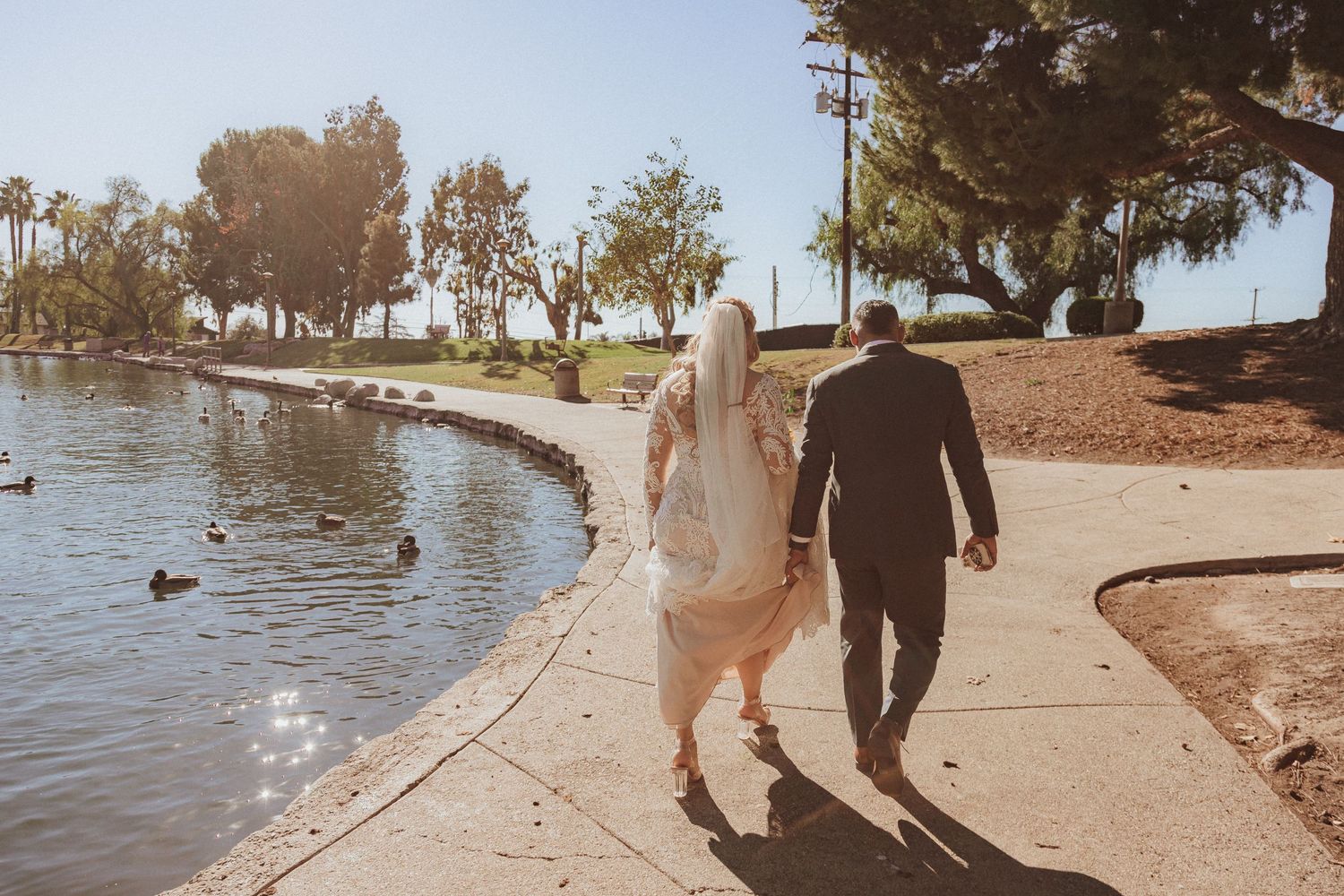 Couple strolls along scenic lakeside path with wedding attire and trailing veil.