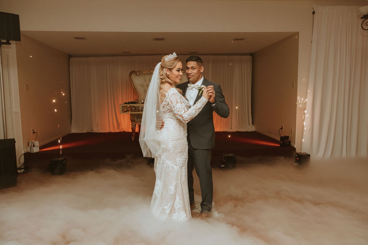 Wedding couple dances together on a fog-covered dance floor with warm lighting and white decor.