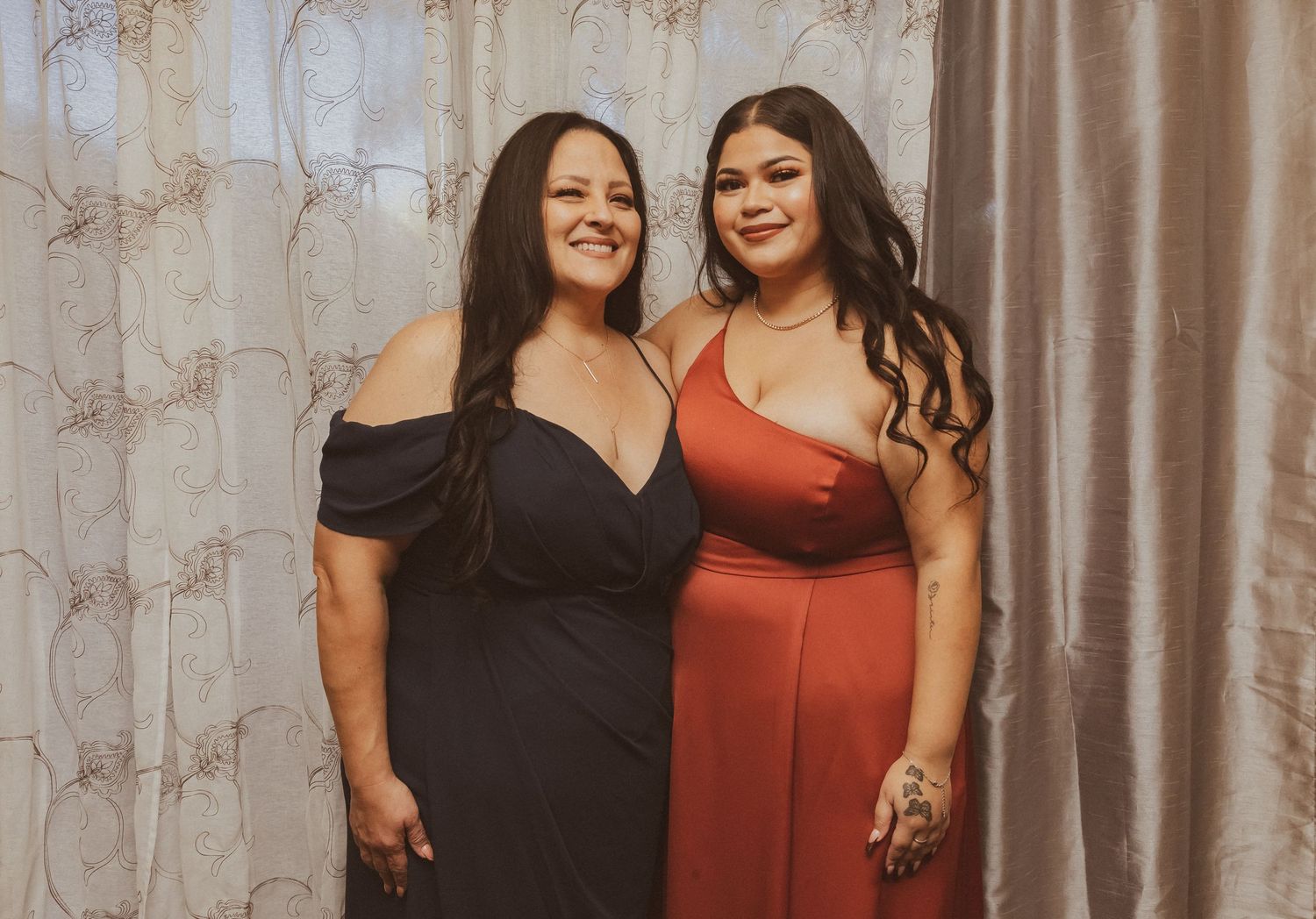 Two women pose together wearing elegant off-shoulder evening gowns in red and black.