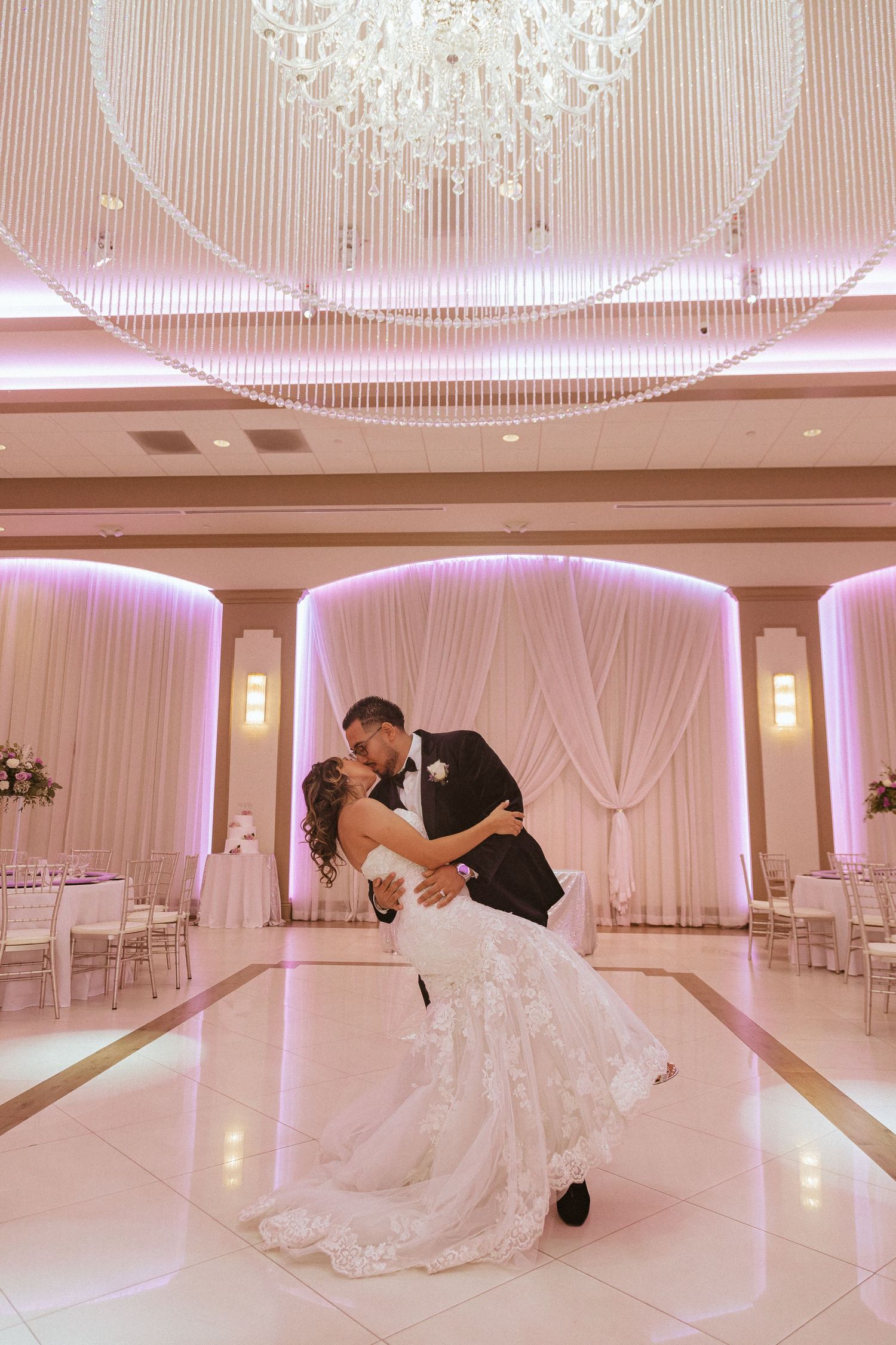 Romantic dip pose under crystal chandelier in elegant ballroom with pink uplighting and white draping.