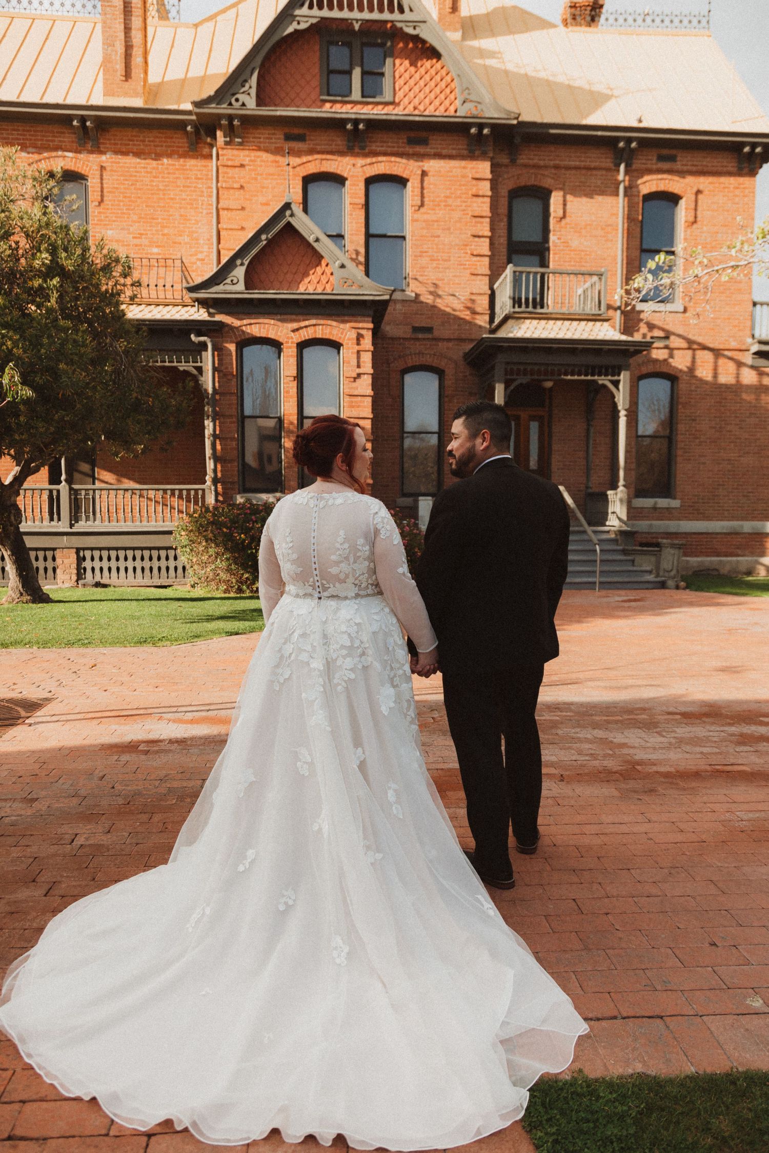 A couple in wedding attire walk towards a historic brick building with ornate Victorian architecture.