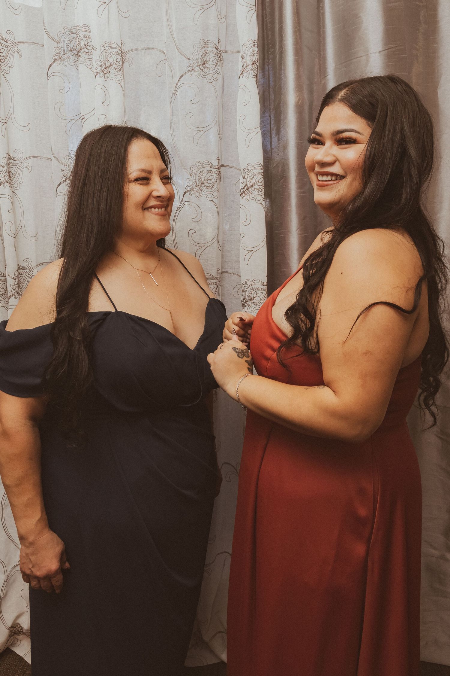 Women wearing black and burgundy formal dresses share a happy moment against a curtained backdrop.