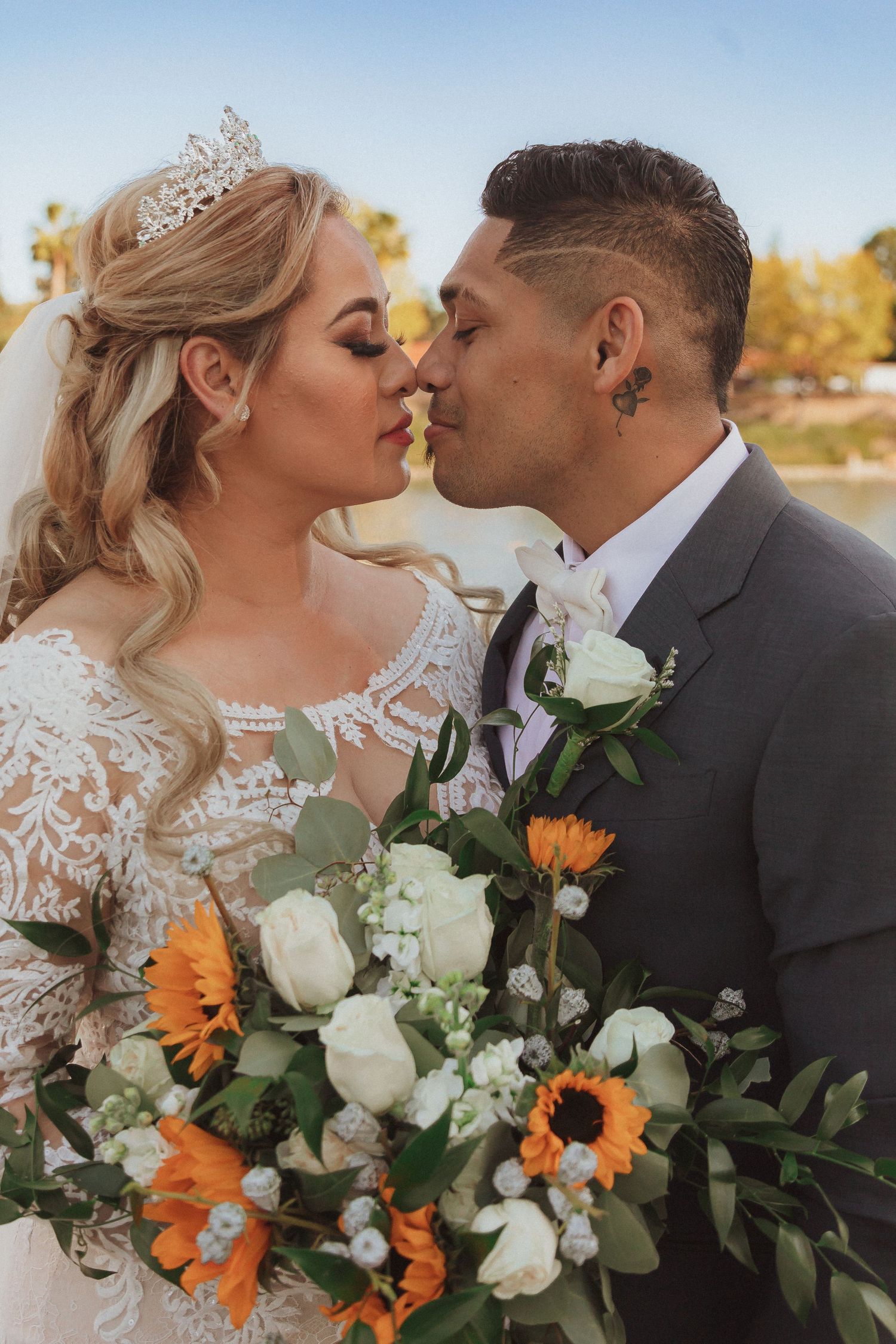 Romantic wedding portrait by the lake with bride holding vibrant sunflower and white rose bouquet.