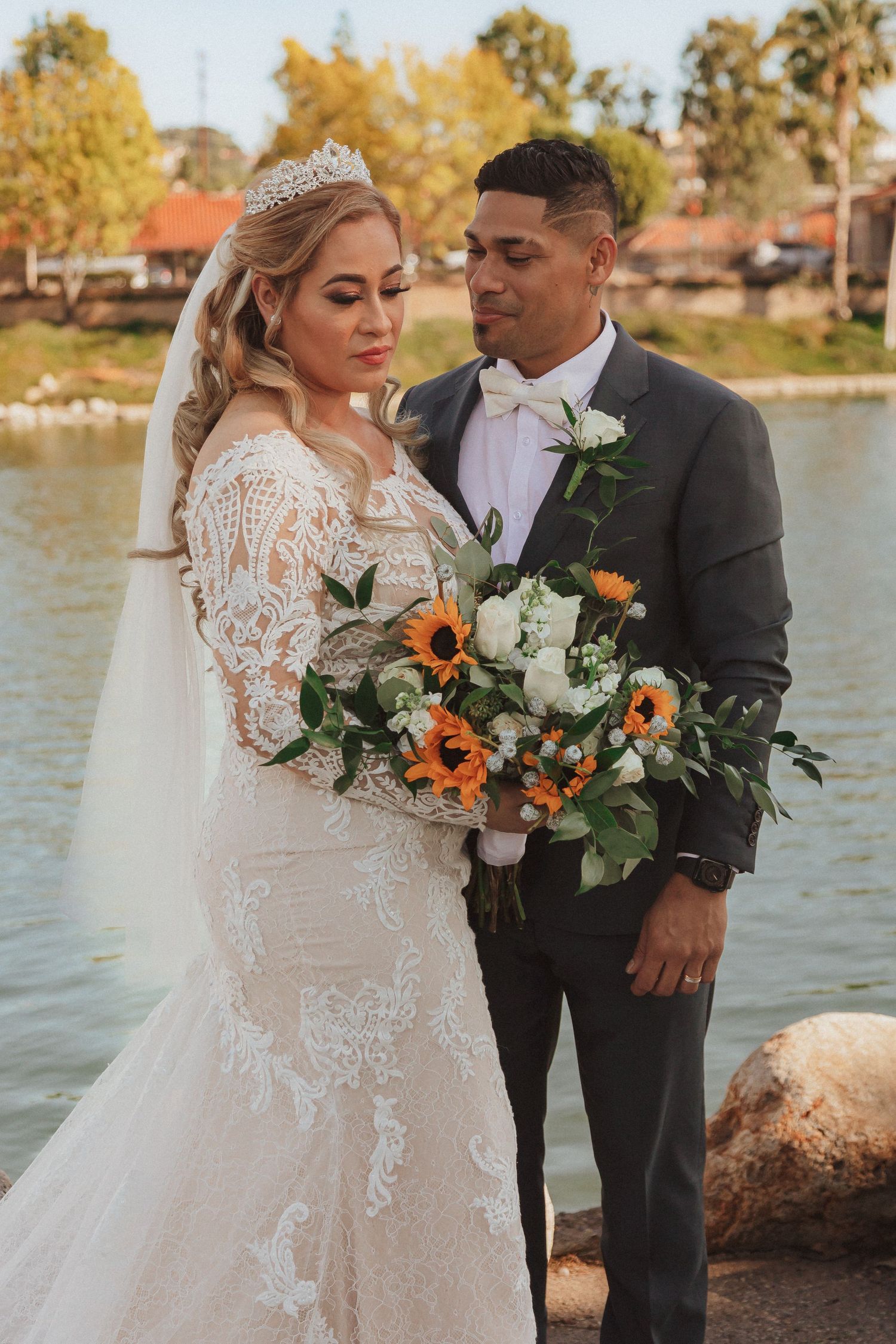Wedding couple poses by a lake with a stunning fall bouquet featuring orange and white flowers.