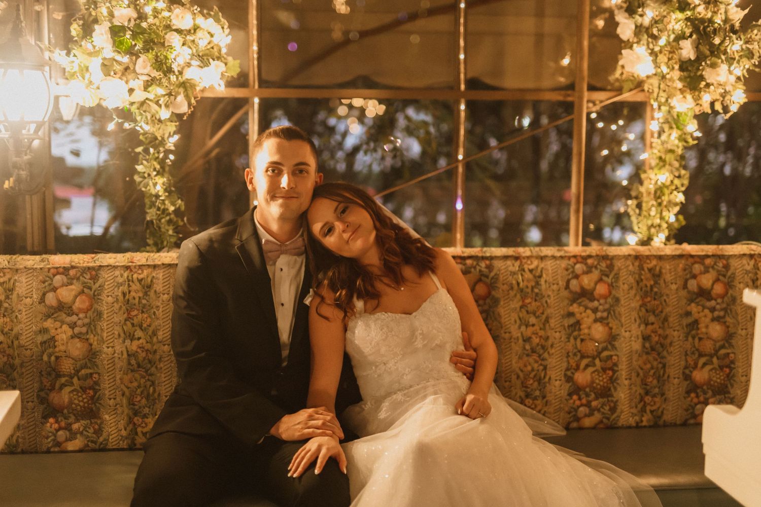 Romantic evening portrait of newlyweds sitting together under glowing fairy lights and floral arrangements.