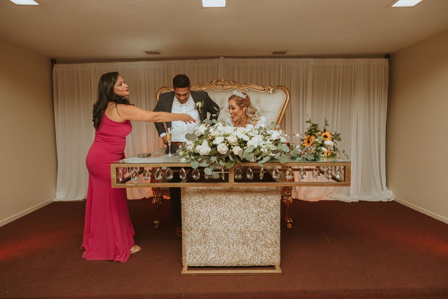 Wedding party celebrating with dance moves behind decorated reception table with white floral arrangements.