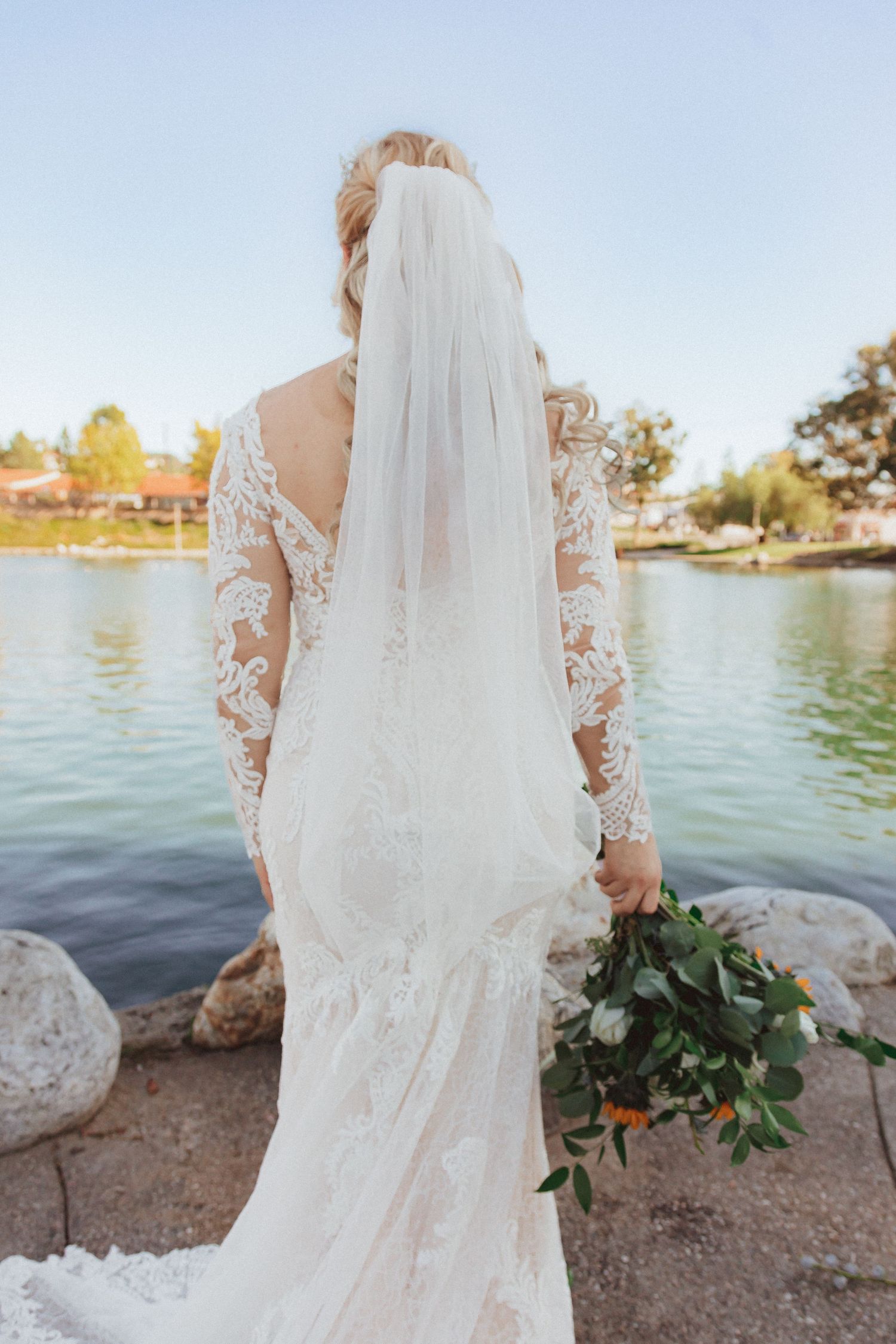 Back view of wedding gown showing detailed lace sleeves and long veil against lake backdrop.