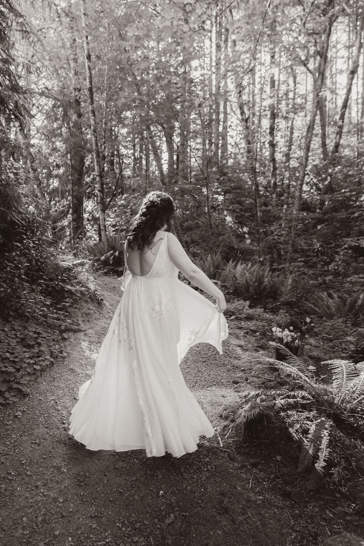 A person in a flowing white wedding dress twirls on a forest path surrounded by trees.
