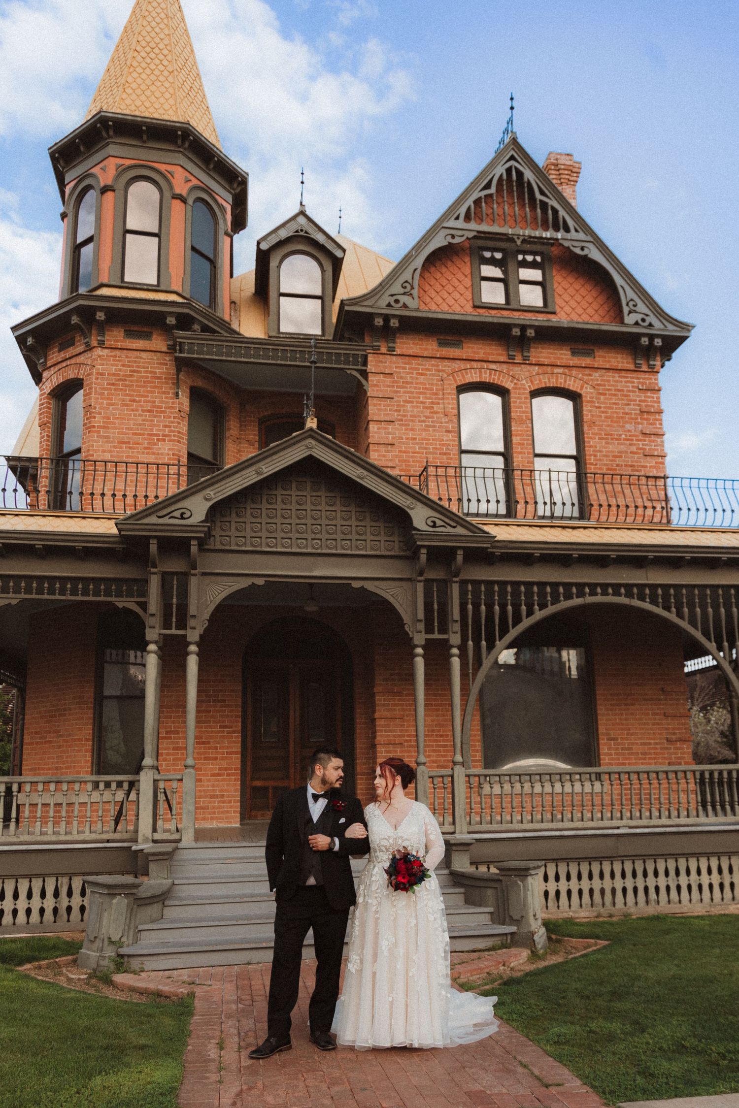 A romantic wedding portrait in front of a grand Victorian mansion with brick facade and decorative trim.
