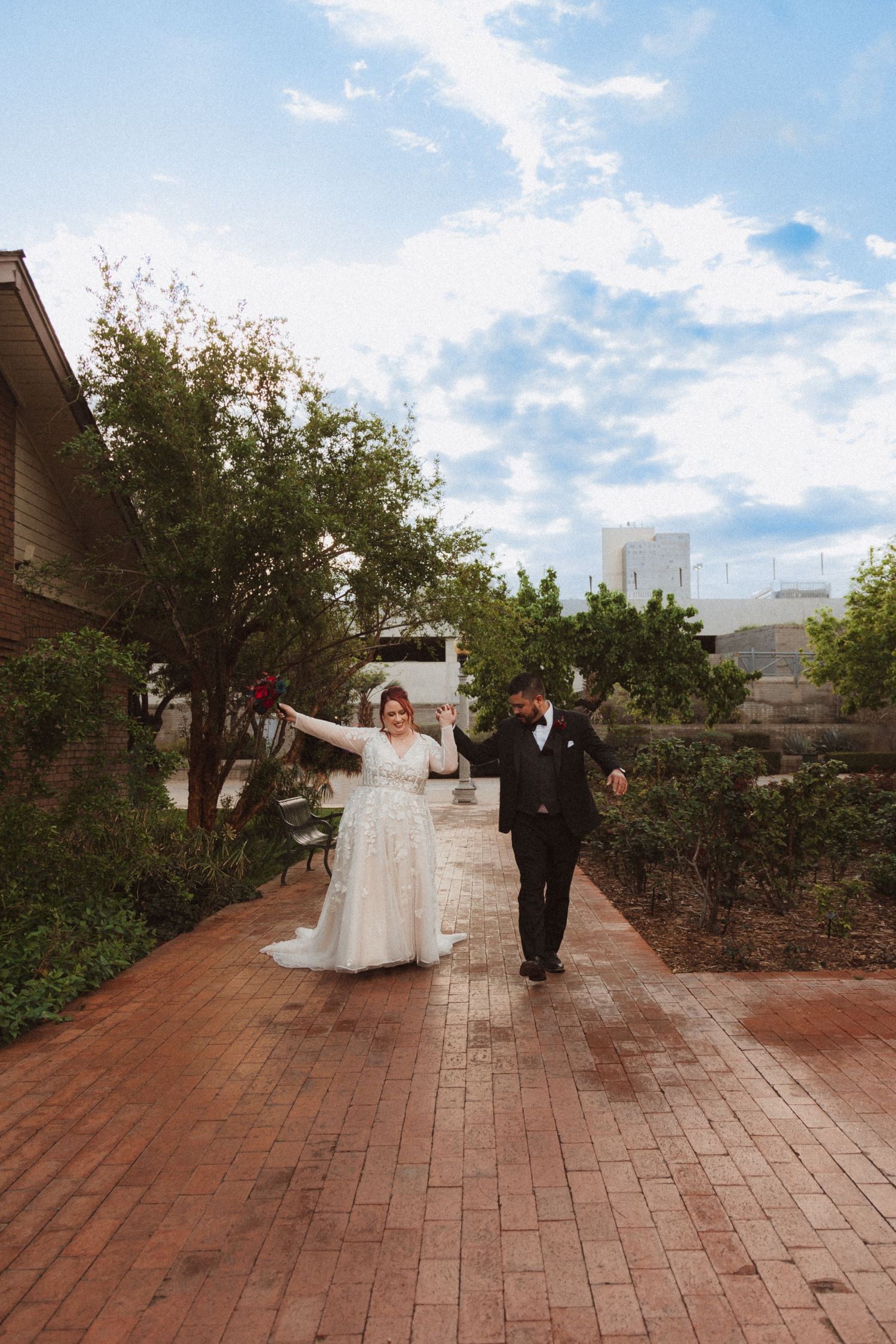 A celebratory wedding moment captured on a brick pathway with arms raised in joy.