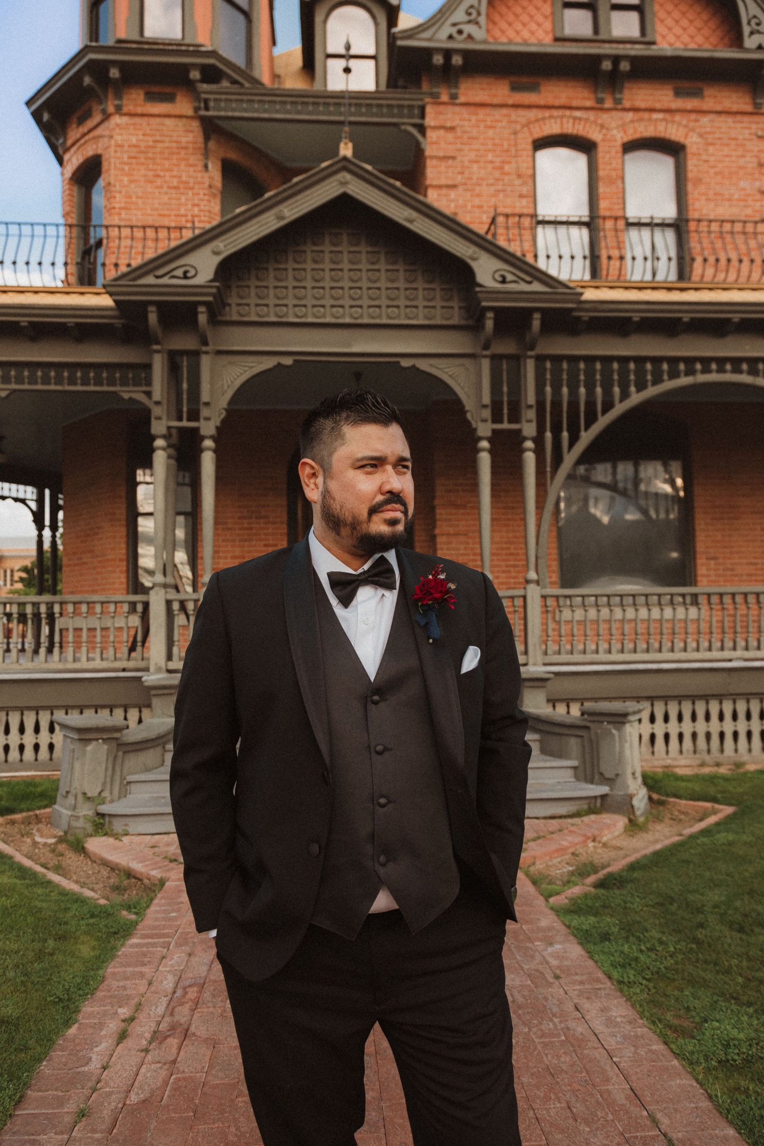 A groom in a black tuxedo poses in front of a Victorian brick mansion with ornate architectural details.