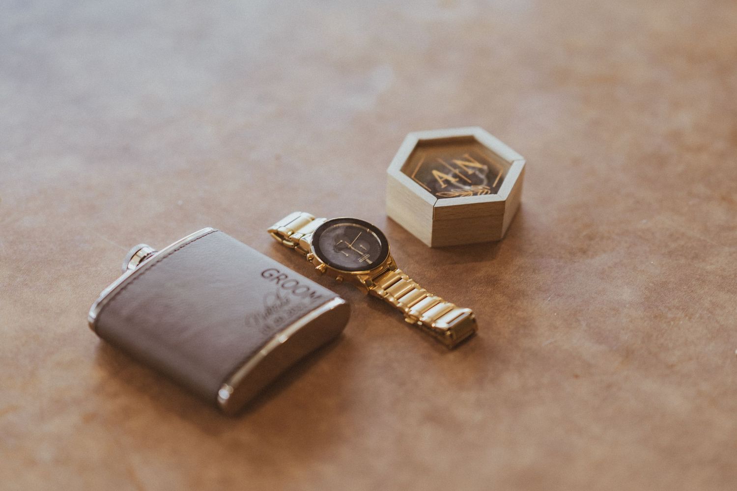 Men's accessories including leather wallet, gold watch and hexagonal box arranged on brown surface.