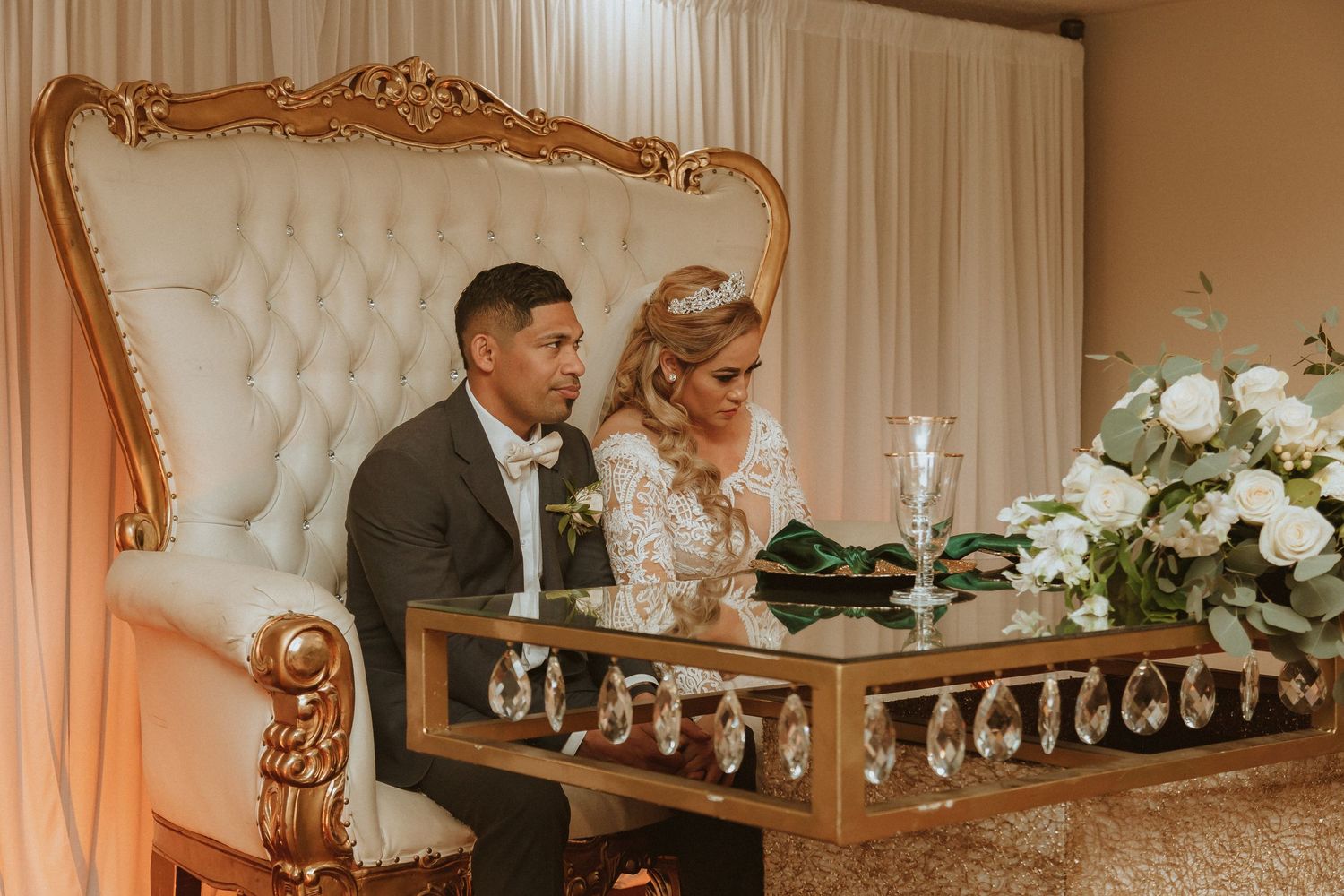 A couple seated at an elegant gold-trimmed sweetheart table with white tufted backing and floral decor.