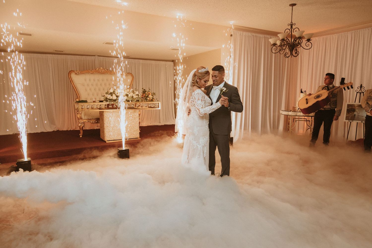 Newlyweds dance together surrounded by dramatic fog effects and cold spark fountains at wedding reception.