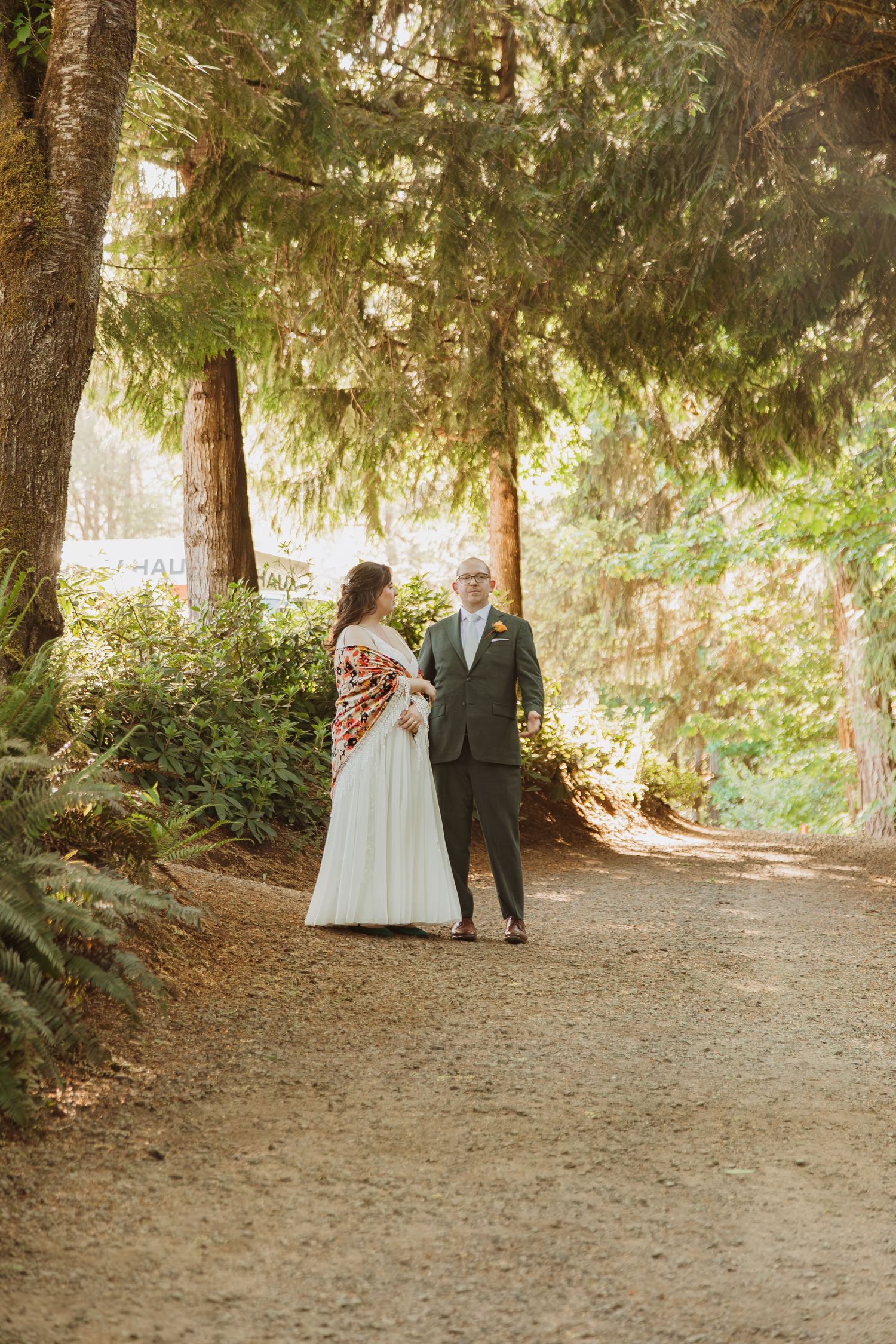 A wedding couple stands together on a tree-lined path dappled with golden sunlight.