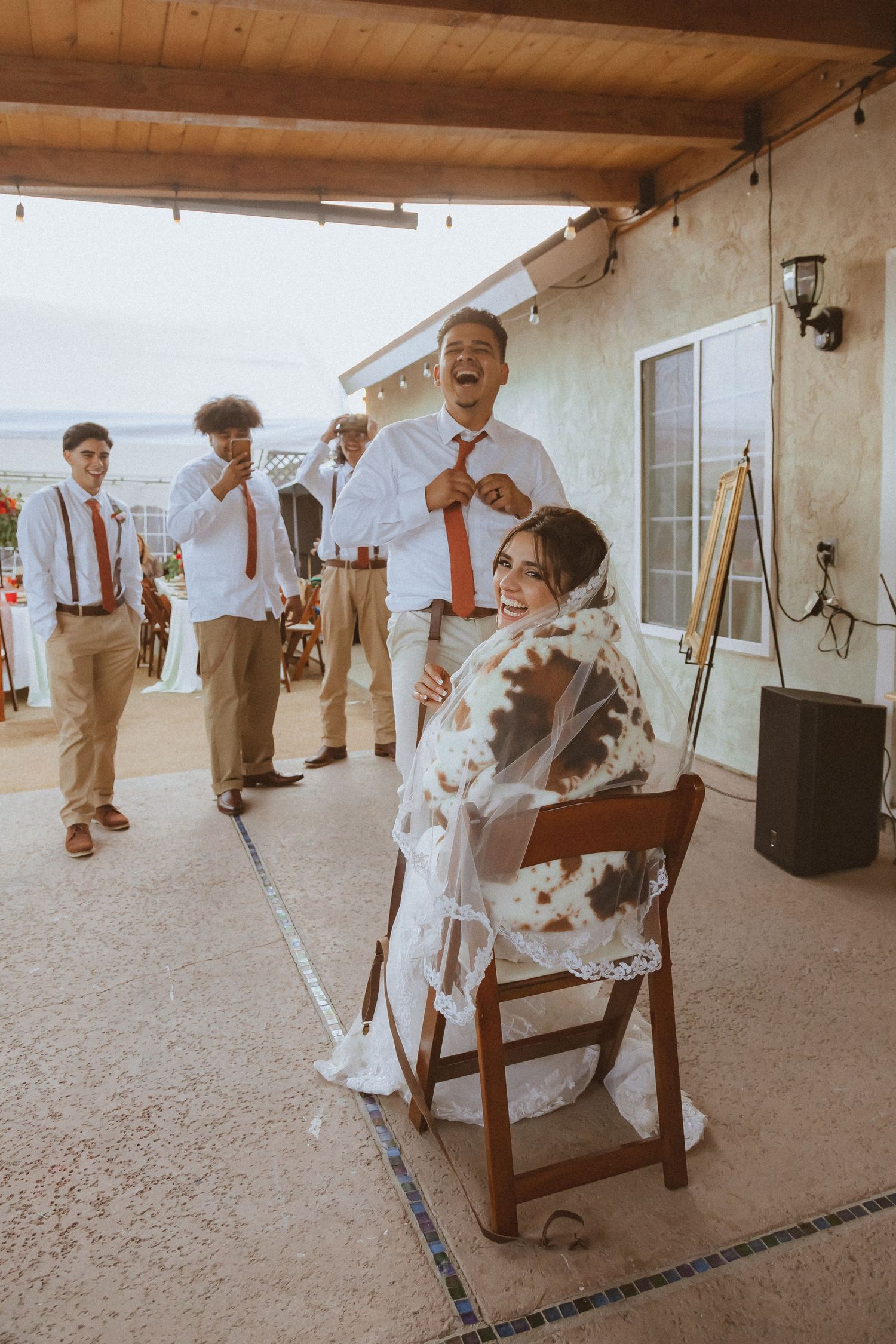 Wedding guests laugh and celebrate as newlyweds share a fun moment on an outdoor patio.