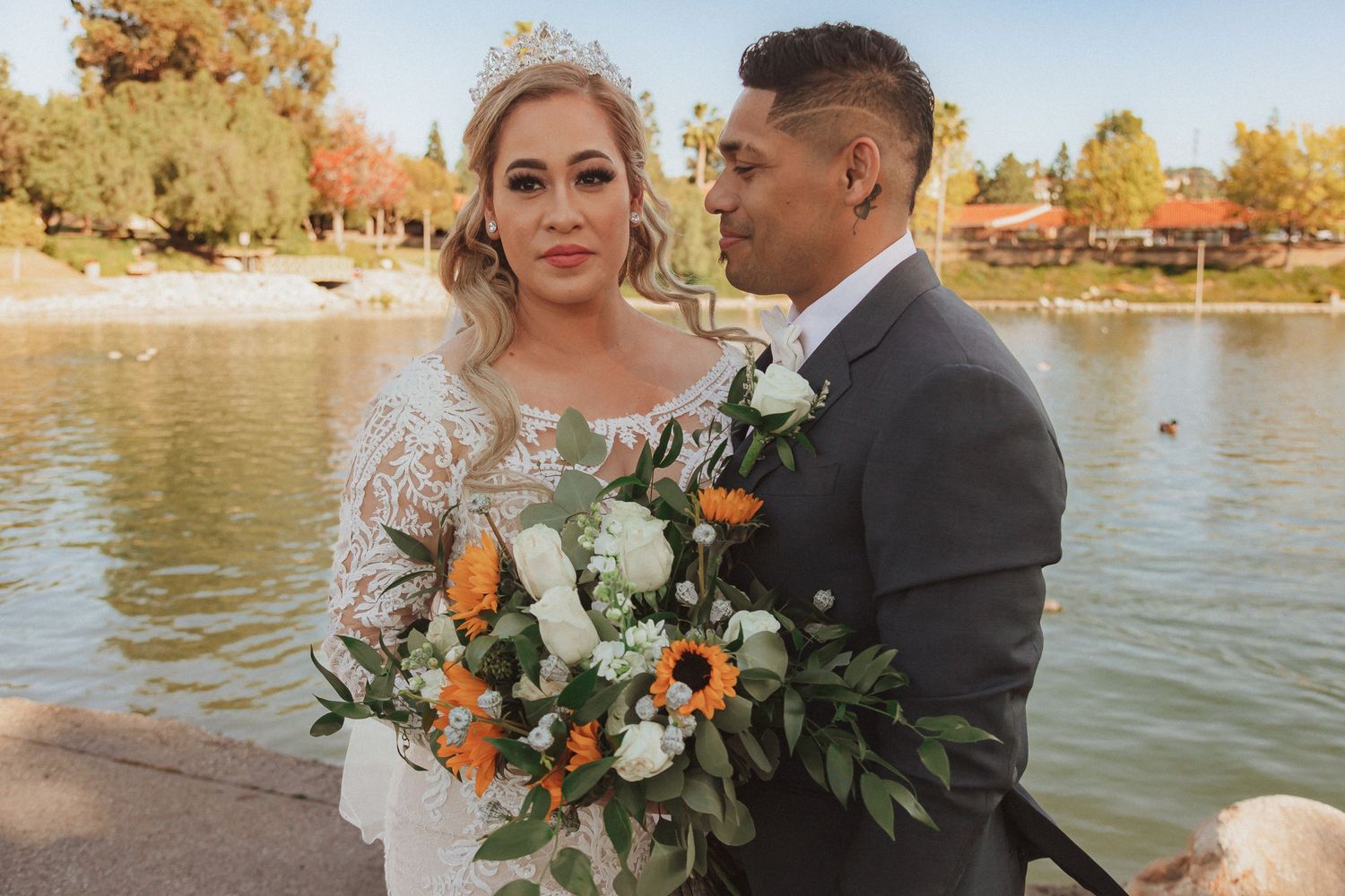 Couple shares intimate moment while holding beautiful orange and white wedding bouquet by lakeside.