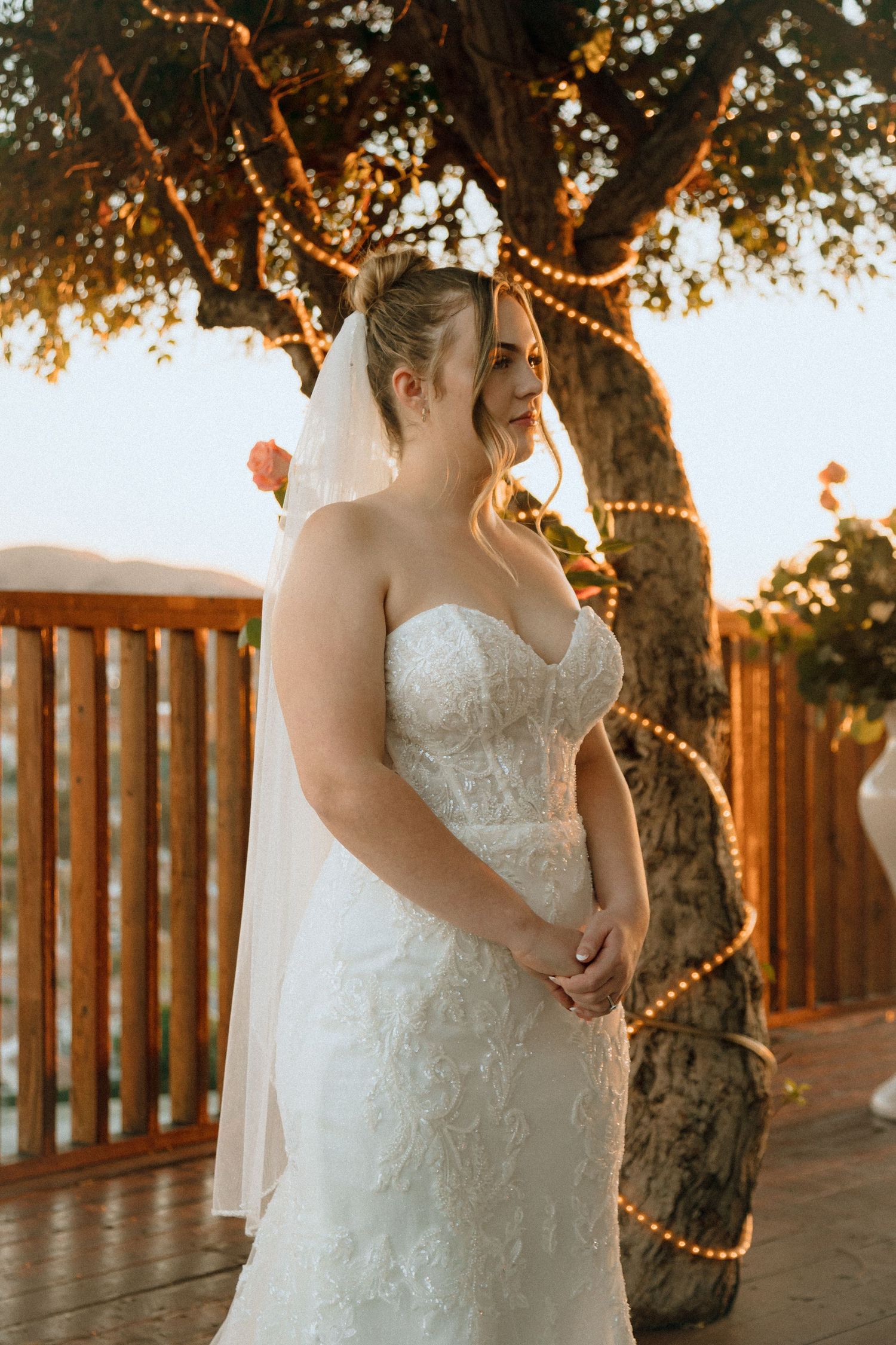 A bride in a strapless lace wedding dress stands on a deck at golden hour with twinkle lights in the trees behind her.