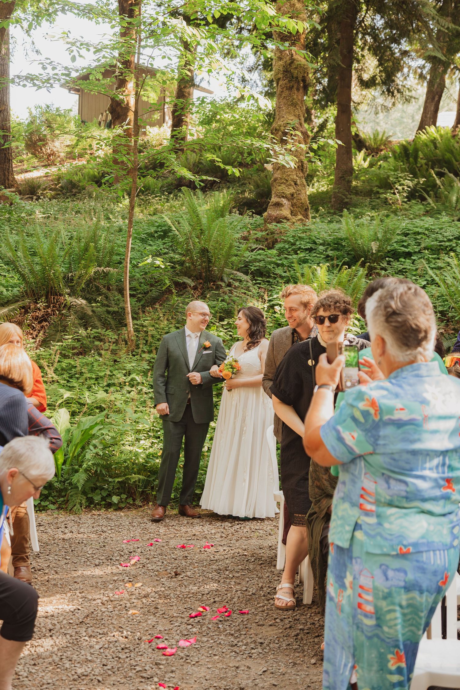 An intimate forest wedding ceremony with guests watching on a gravel path surrounded by ferns.