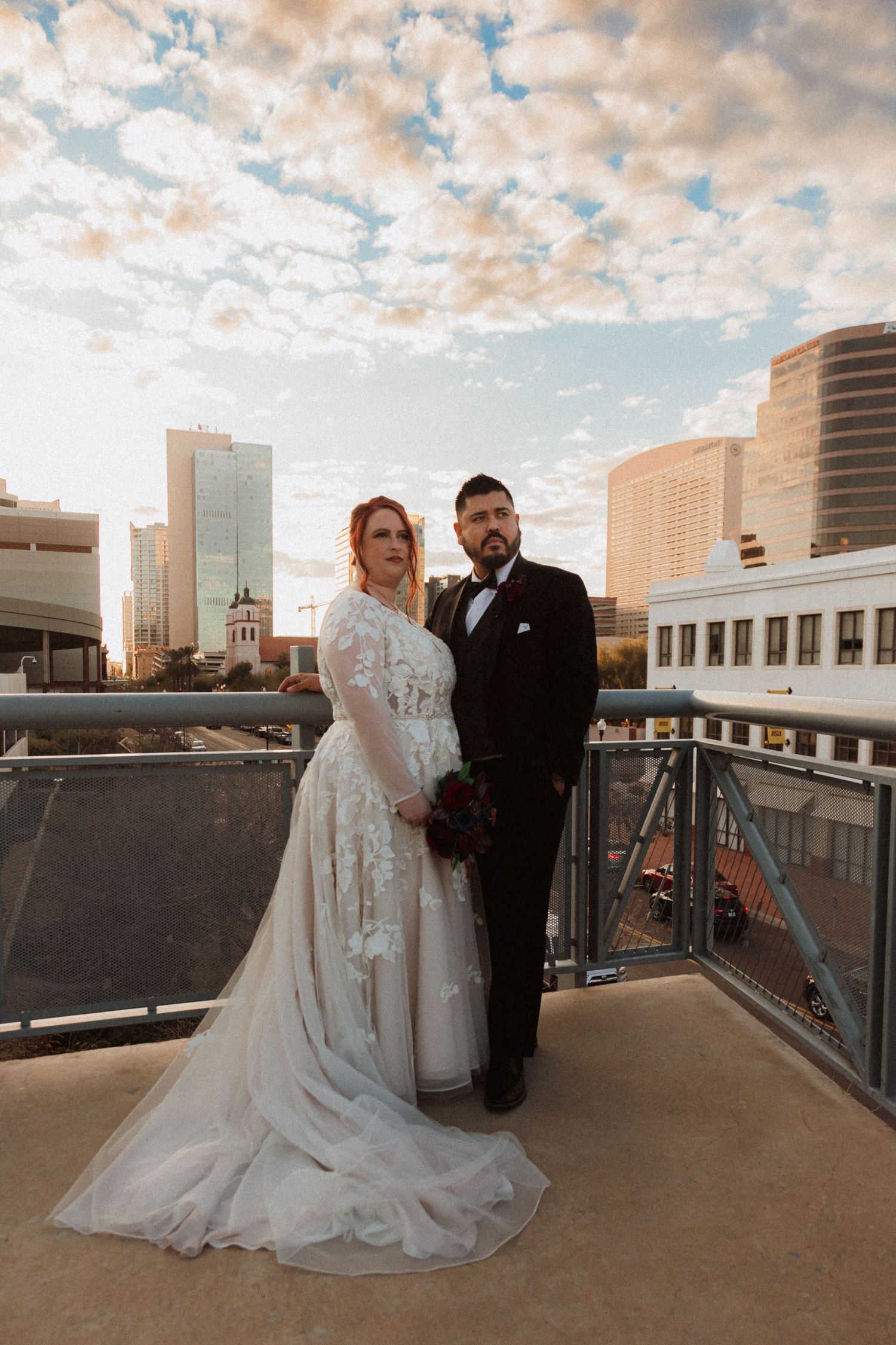 Elegant wedding couple poses on rooftop with urban skyline and dramatic cloudy sunset.