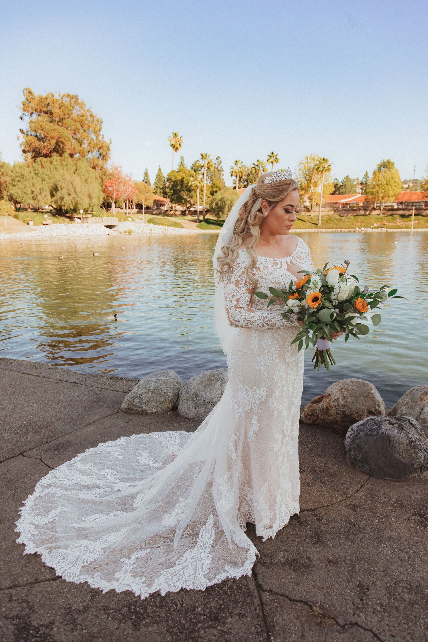Side view of bride in long-sleeved lace wedding dress holding autumn bouquet by water.