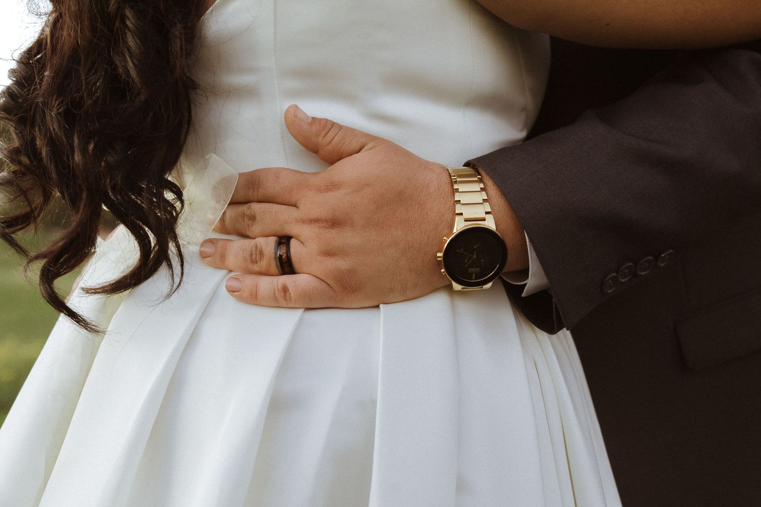 Close-up of couple's hands with wedding rings against white dress and brown suit.